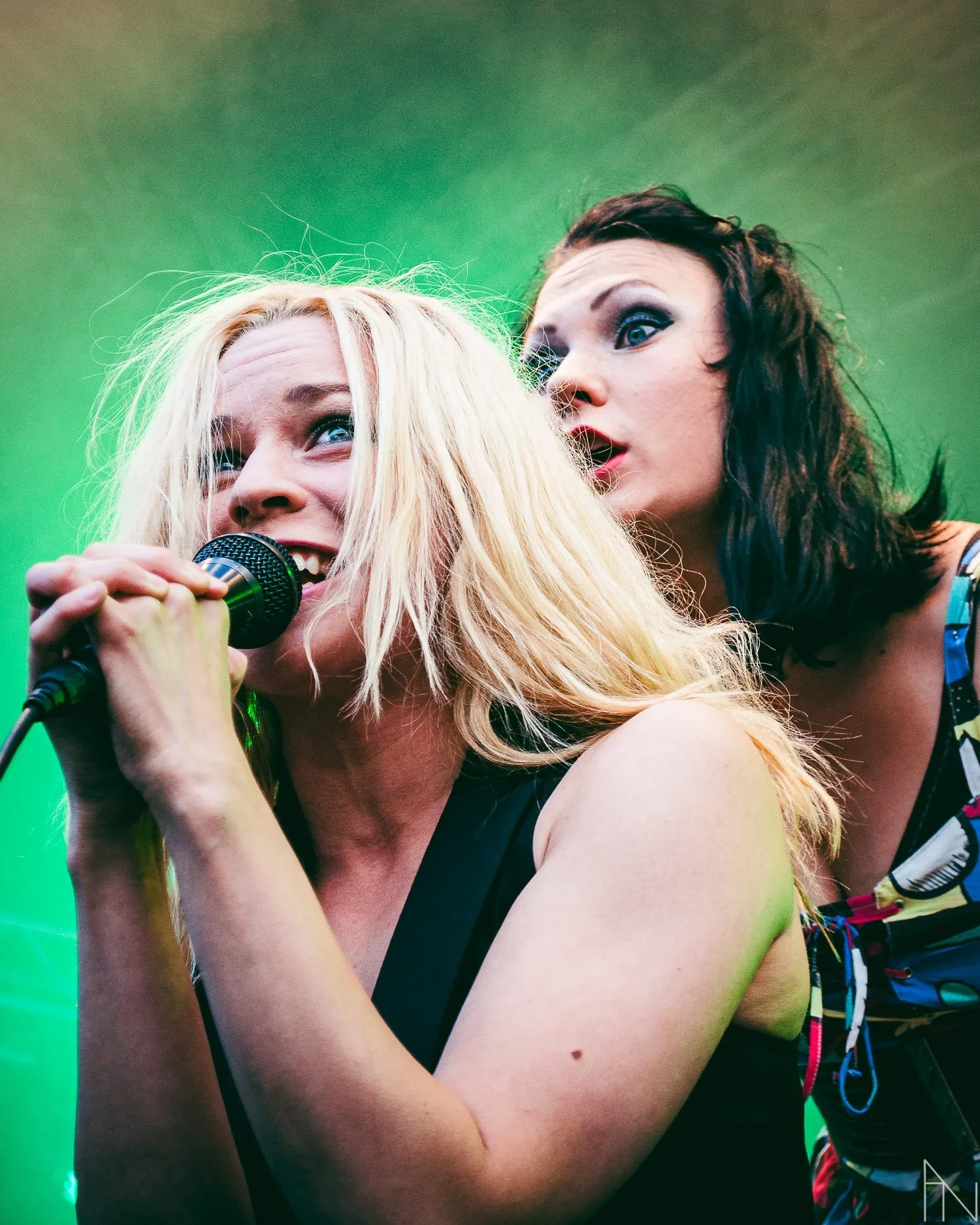 Kaksi naista, toinen laulamassa mikrofonia ja toinen taustalla, esiintymässä lavalla, vihreä valaistus. PMMP Jurassic Rock Mikkeli.