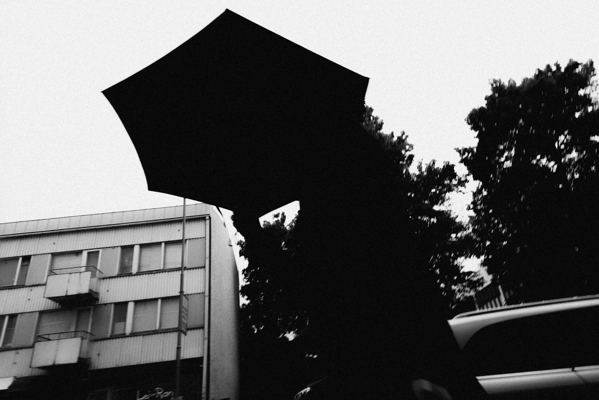 Henkilö pitää mustaa sateenvarjoa ulkona, taustalla rakennus ja puita, mustavalkoinen kuva.
