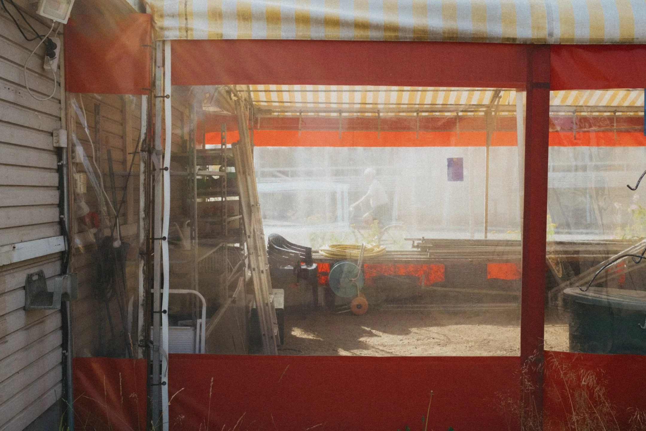 Kuvassa varjostettu ulkotila, jossa on muutamia työkaluja ja rakennusmateriaalit, näkyy myös henkilö polkupyörällä taustalla.