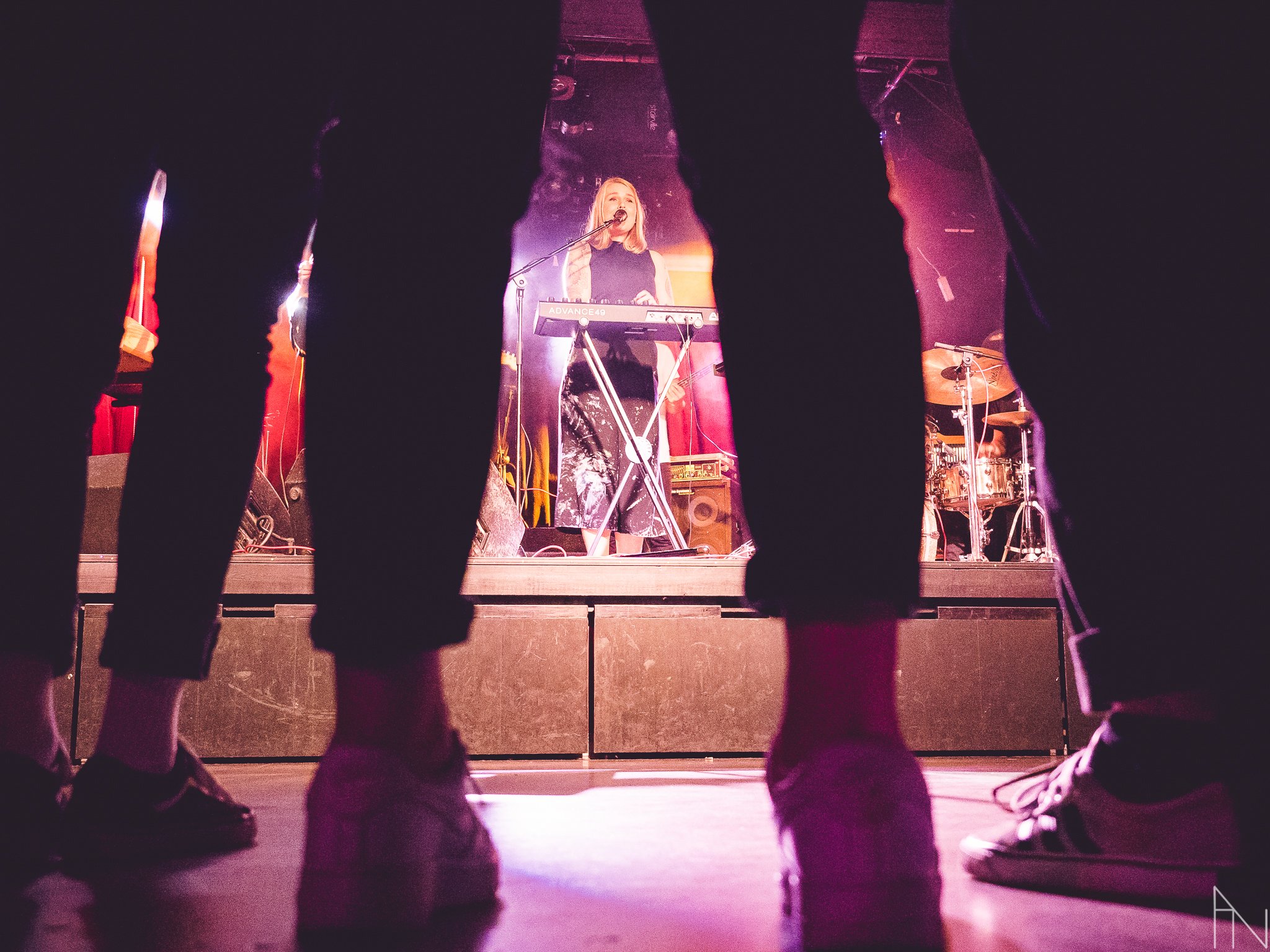 Musiikki keikalla, nainen soittaa kosketinsoitinta ja laulaa taustavalojen alla, näkyvät myös rummut. Etualalla yleisön jalkoja.