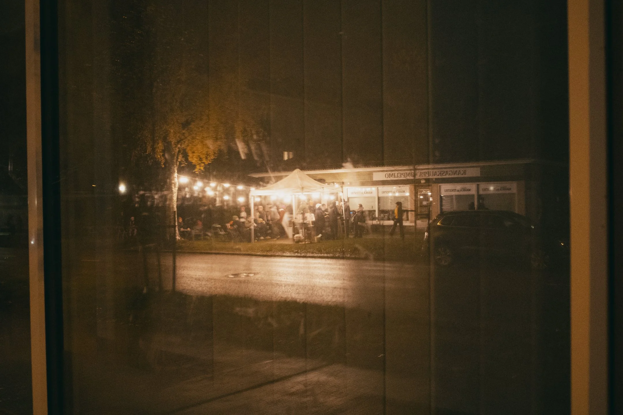 Ilta- tai yökuva ravintolasta tai kahvilasta, seen hehkuvilla valoilla ja ihmisillä sisällä ja ulkona.