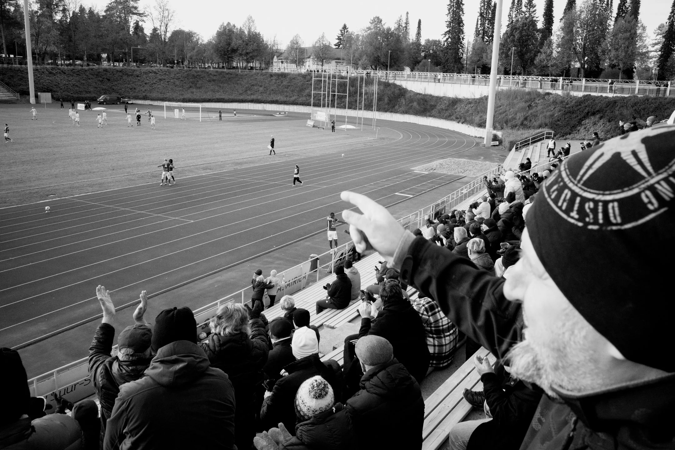 Mustavalkoinen kuva jalkapallo-ottelusta stadionilla, jossa katsomossa ihmiset katsovat peliä, osa niistä juttelee ja jotkut kuvaavat, taustalla näkyy pelaajia kentällä.