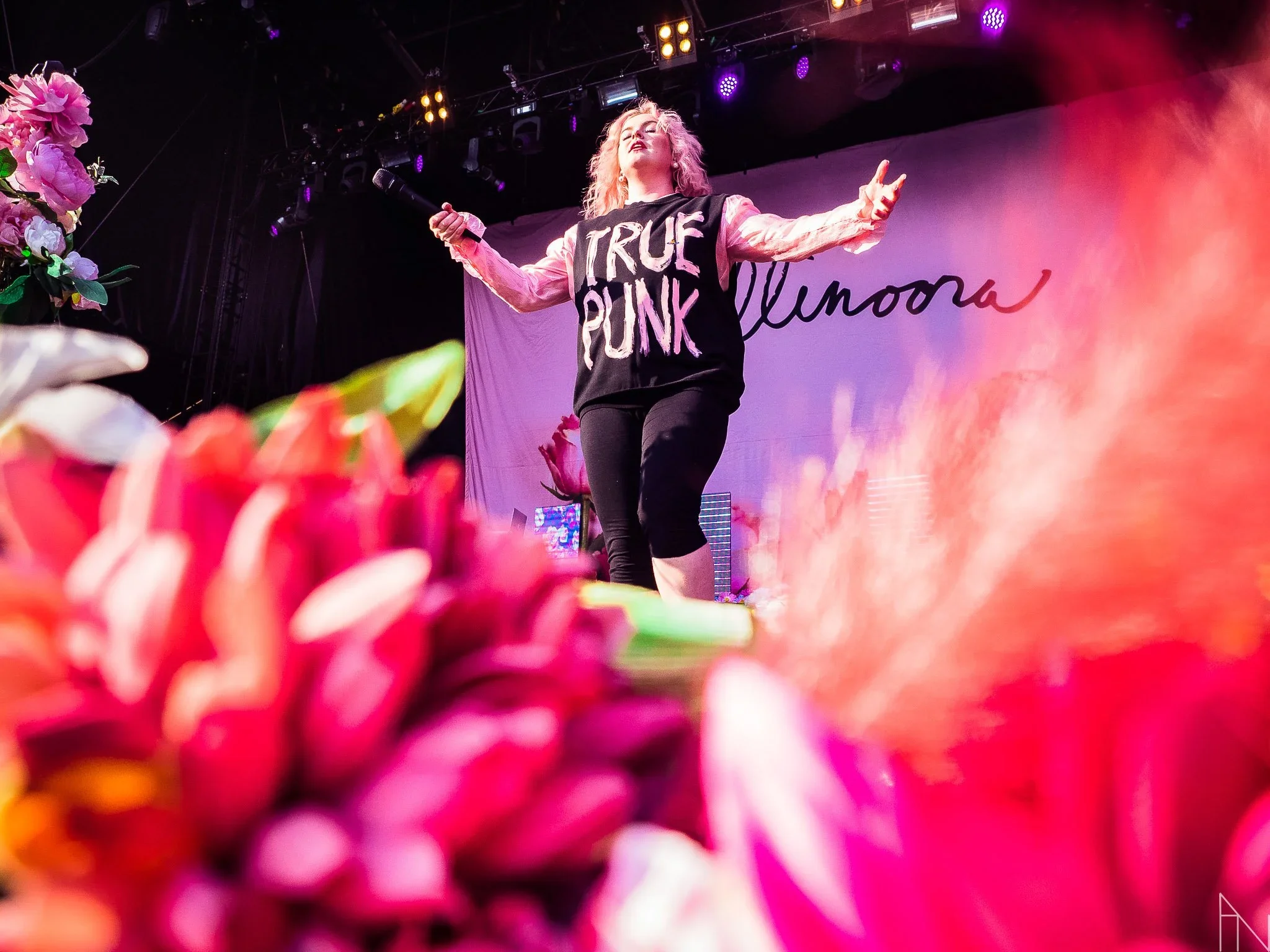 Nainen esiintyy lavalla, jossa on taustalla teksti 'Ellinoora'. Hänellä on päässään vaaleat hiukset ja hän pitää mikrofonia kädessään. Edessä on paljon värikkäitä kukkia, osa näkyy epätarkasti.