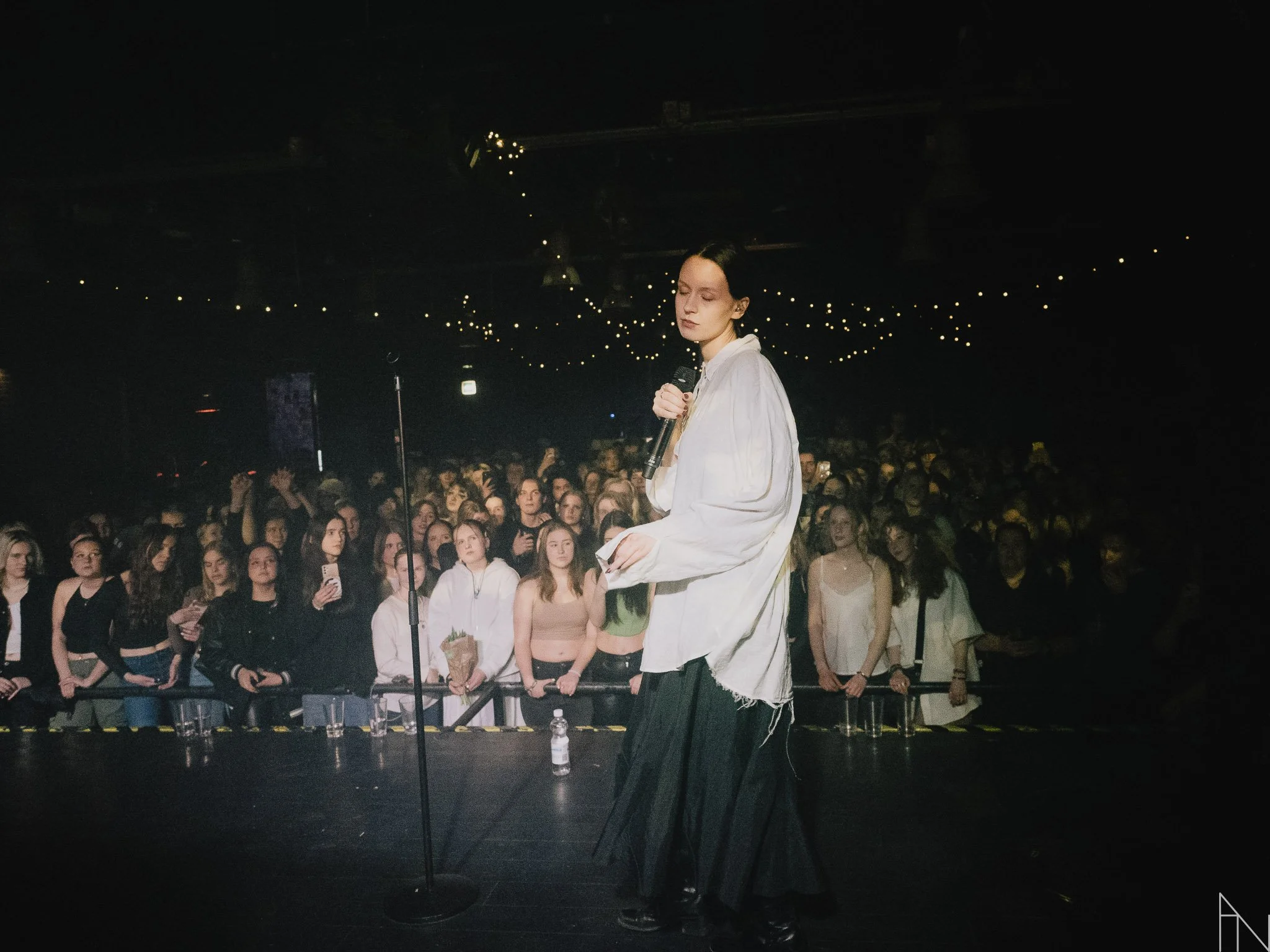 Nainen esiintyy mikrofonin kanssa lavalla, taustalla suuri yleisö ja seinävaloissa pieniä valoja. Pehmoaino Lutakko 2022.