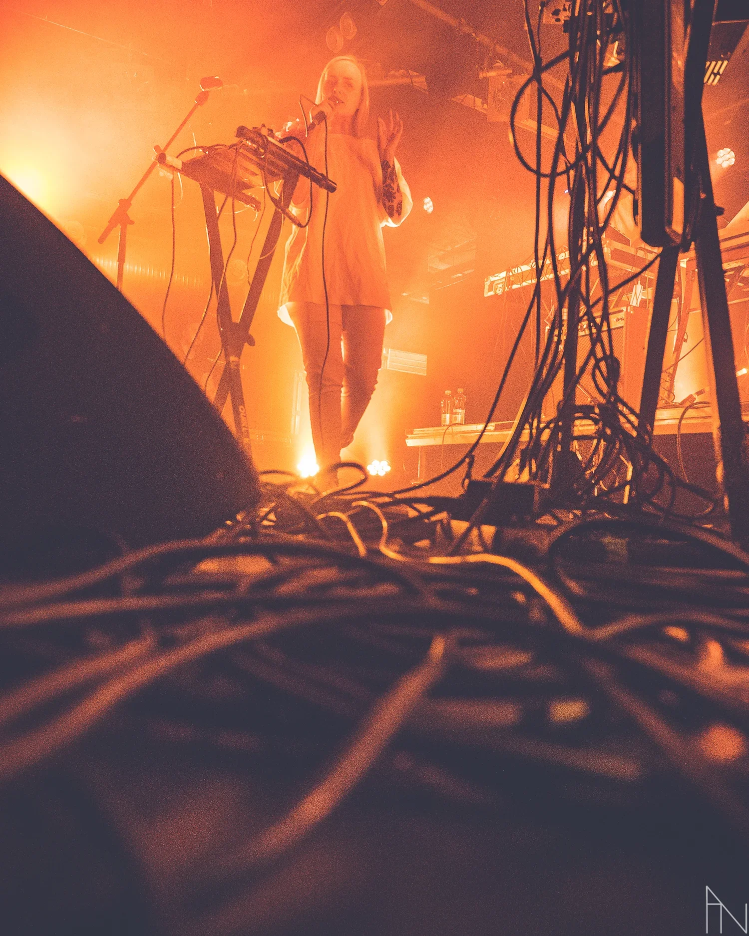 Naisartisti, Ringa Manner, laulaa mikrofoniin lavalla orkesterin taustalla, edessä ja oikealla paljon roikkuvia johtoja. Oranssi kuva.