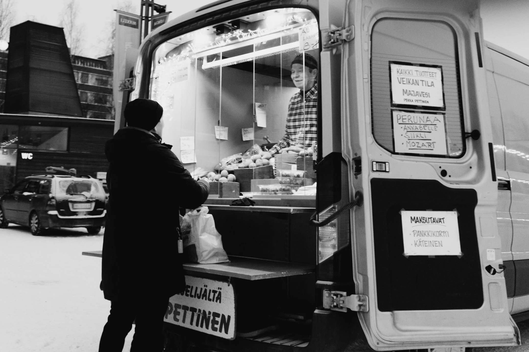 Mies ostaa kasviksia siirtyvästä myyntikojusta ulkona, takana pysäköityjä autoja ja rakennuksia ja loskaa. Mikkelin tori.