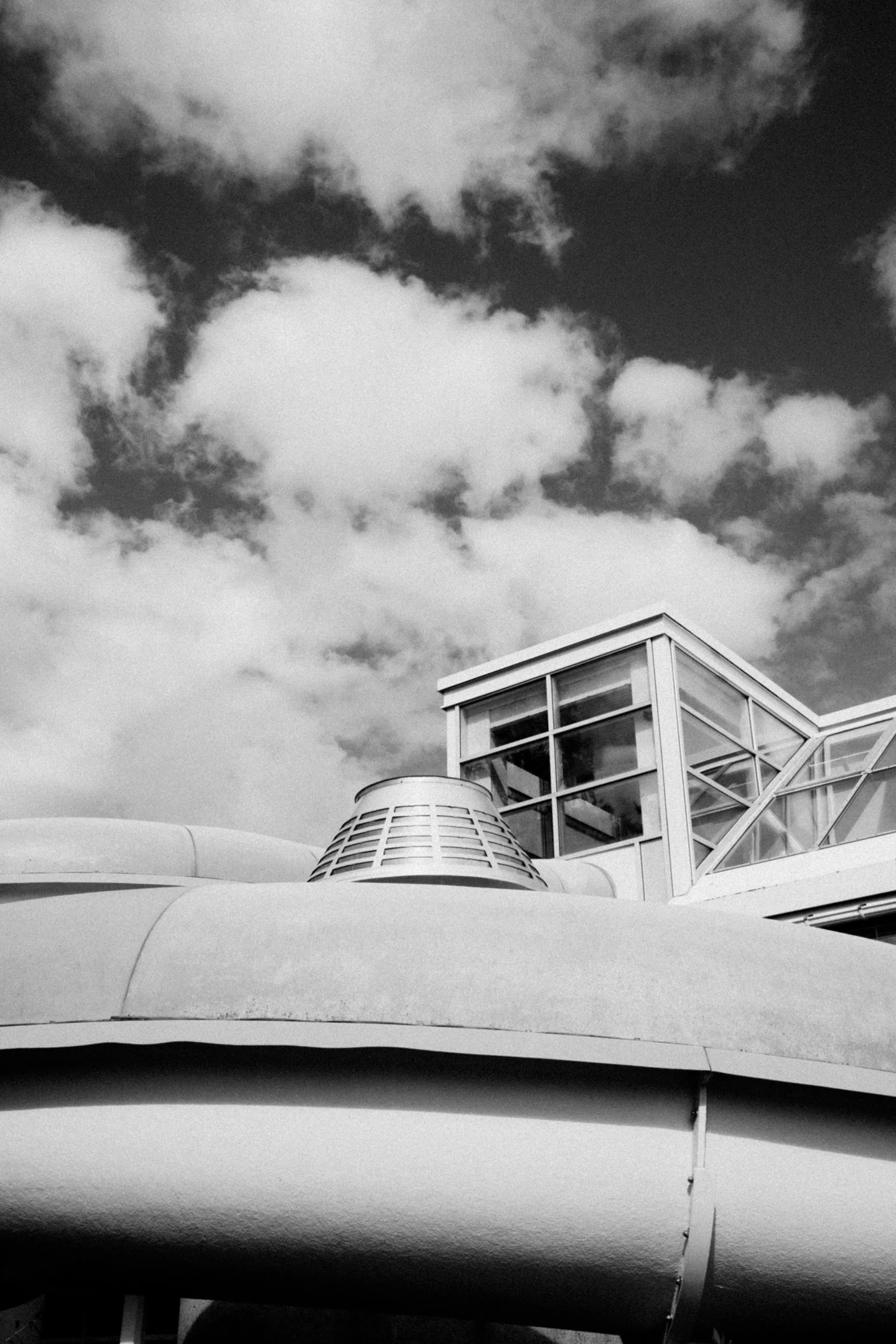 Mustavalkoinen kuva nykyaikaisesta rakennuksesta, jossa on lasi-ikkunoita ja metallirakenteita, taivaalla pilviä.