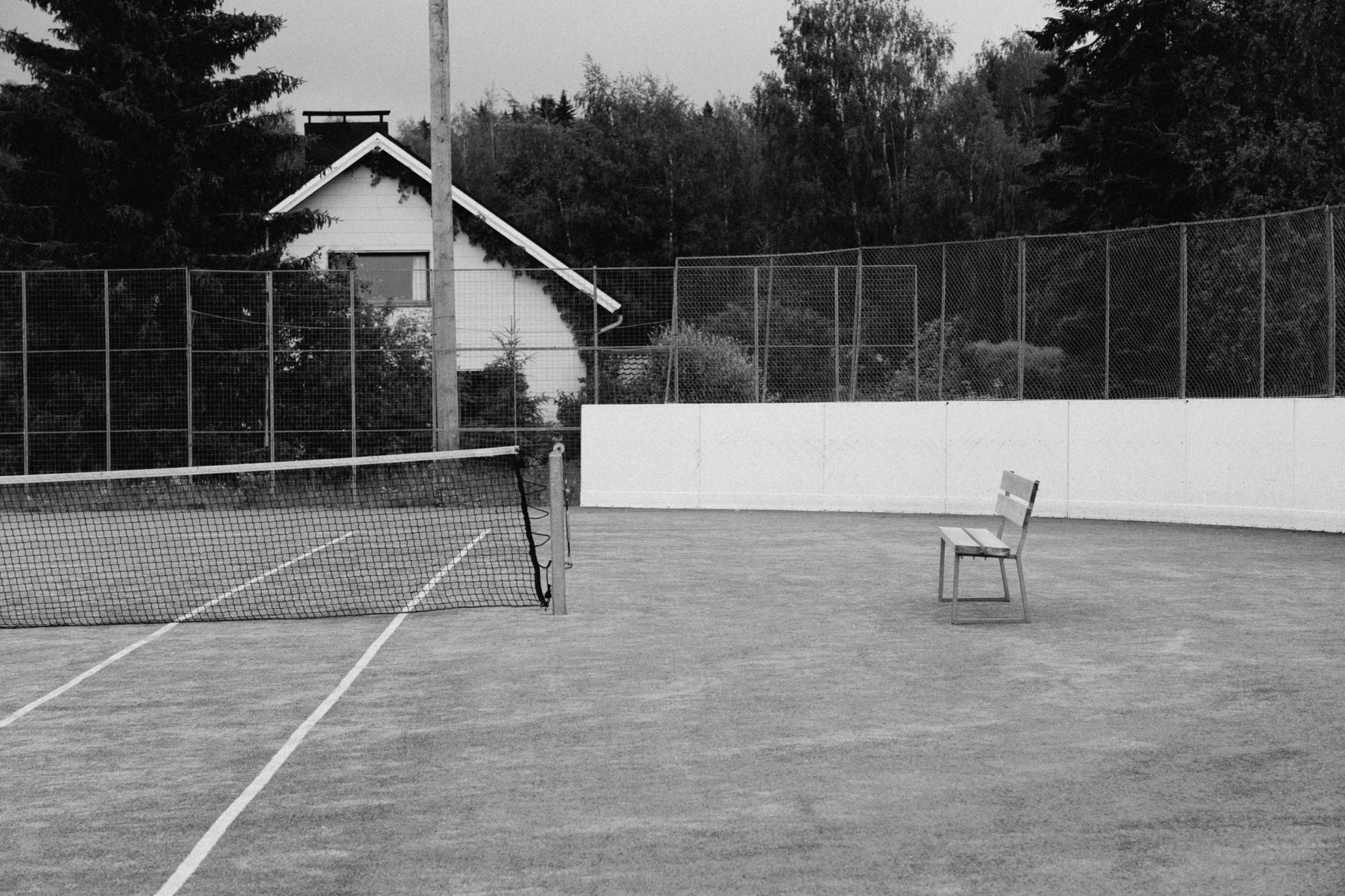 Tenniskenttä, jossa on ristikkoverkko ja puinen penkki, maasto ja pihapiiri taustalla.
