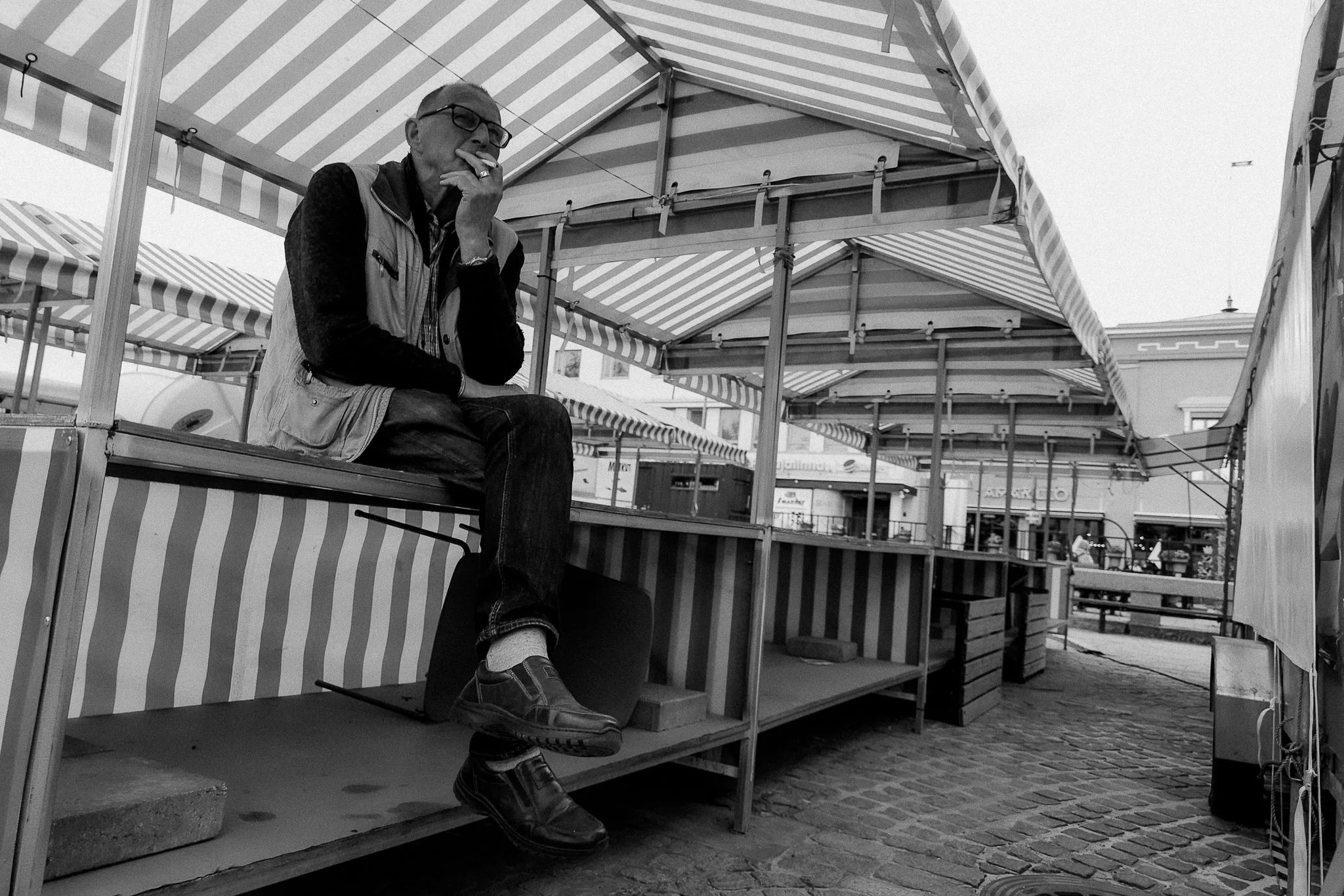 Mies istuu markkinoiden kojuissa, kalteva katto ja paikalleen asetellut myyntiastiat ympärillään, ja hän sytyttää savukkeen.