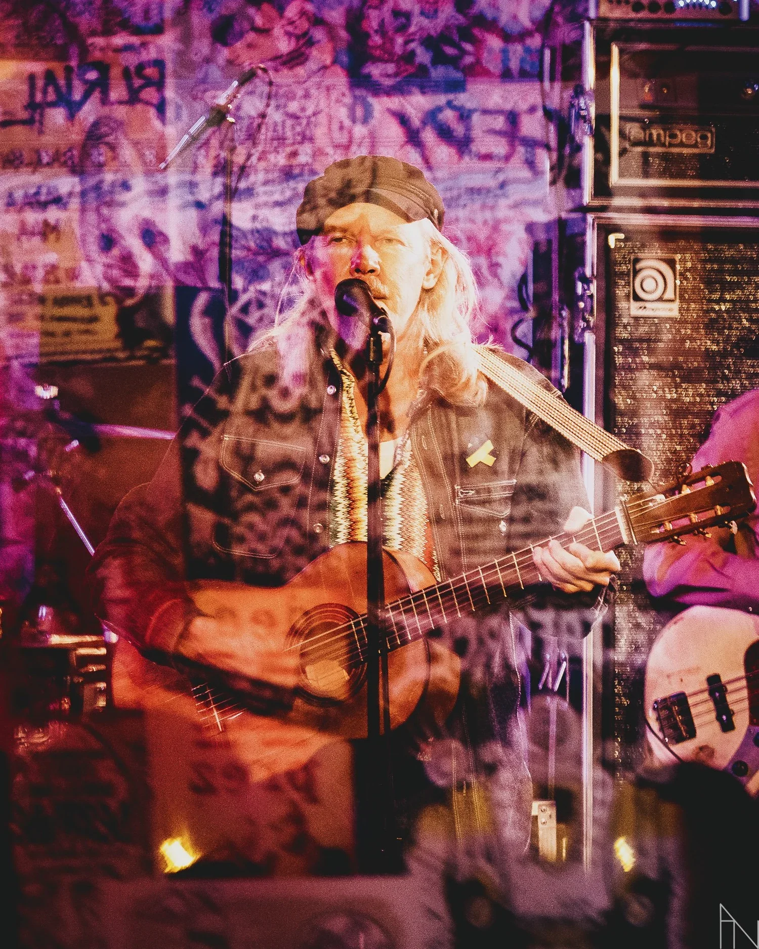 J. Karjalainen soittaa kitaraa ja laulaa mikrofonin ääressä esiintymislavalla, taustalla seinällä graffititaidetta ja musiikkioptiikoita.