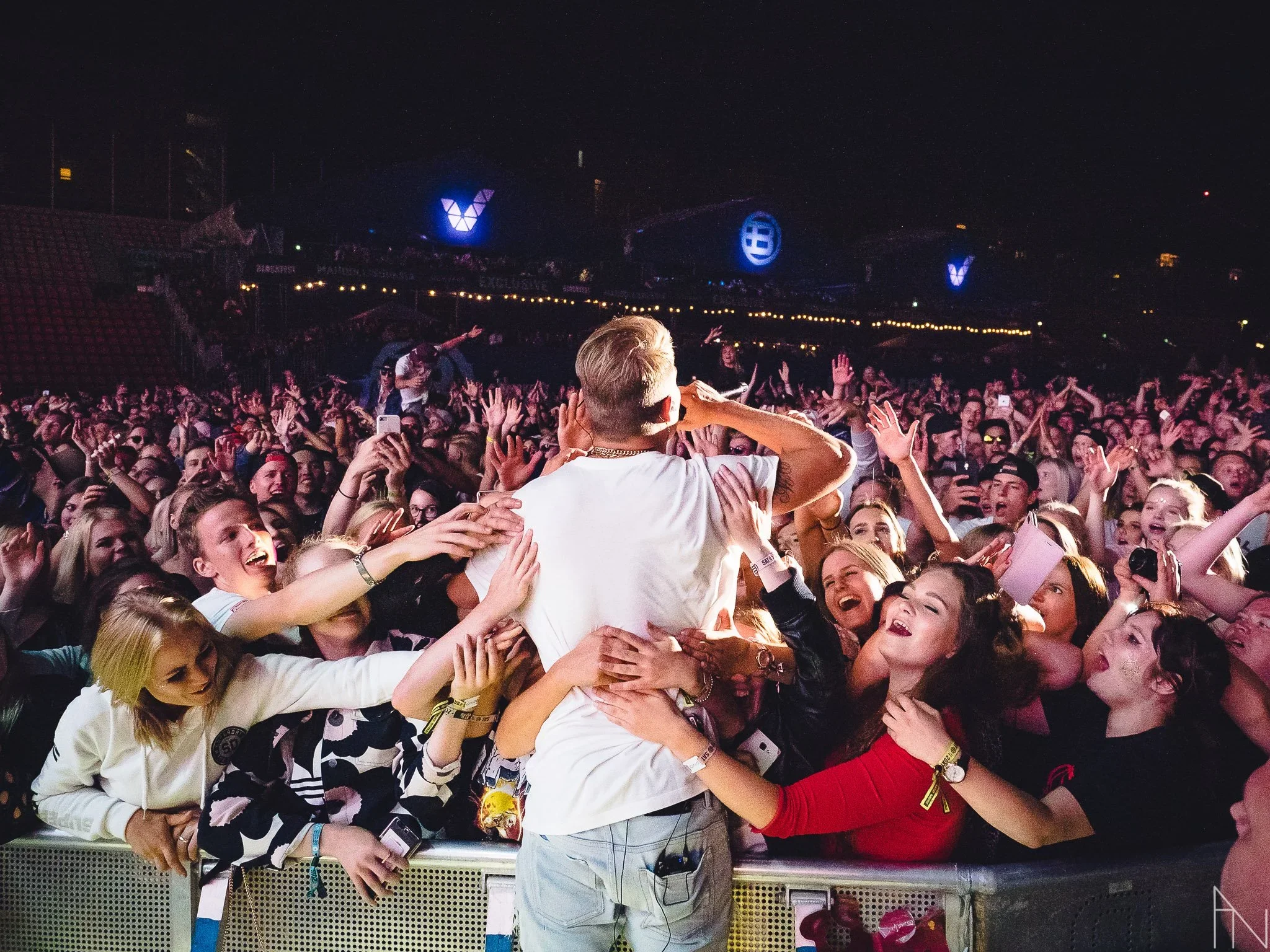 Konsertissa oleva yleisö ja esiintyjä, jossa monia ihmisiä hymyilemässä ja ottamassa valokuvia tai videoita, ilmaisena ja juhlallisena tunnelmana. Yleisö halaa Cheekiä.