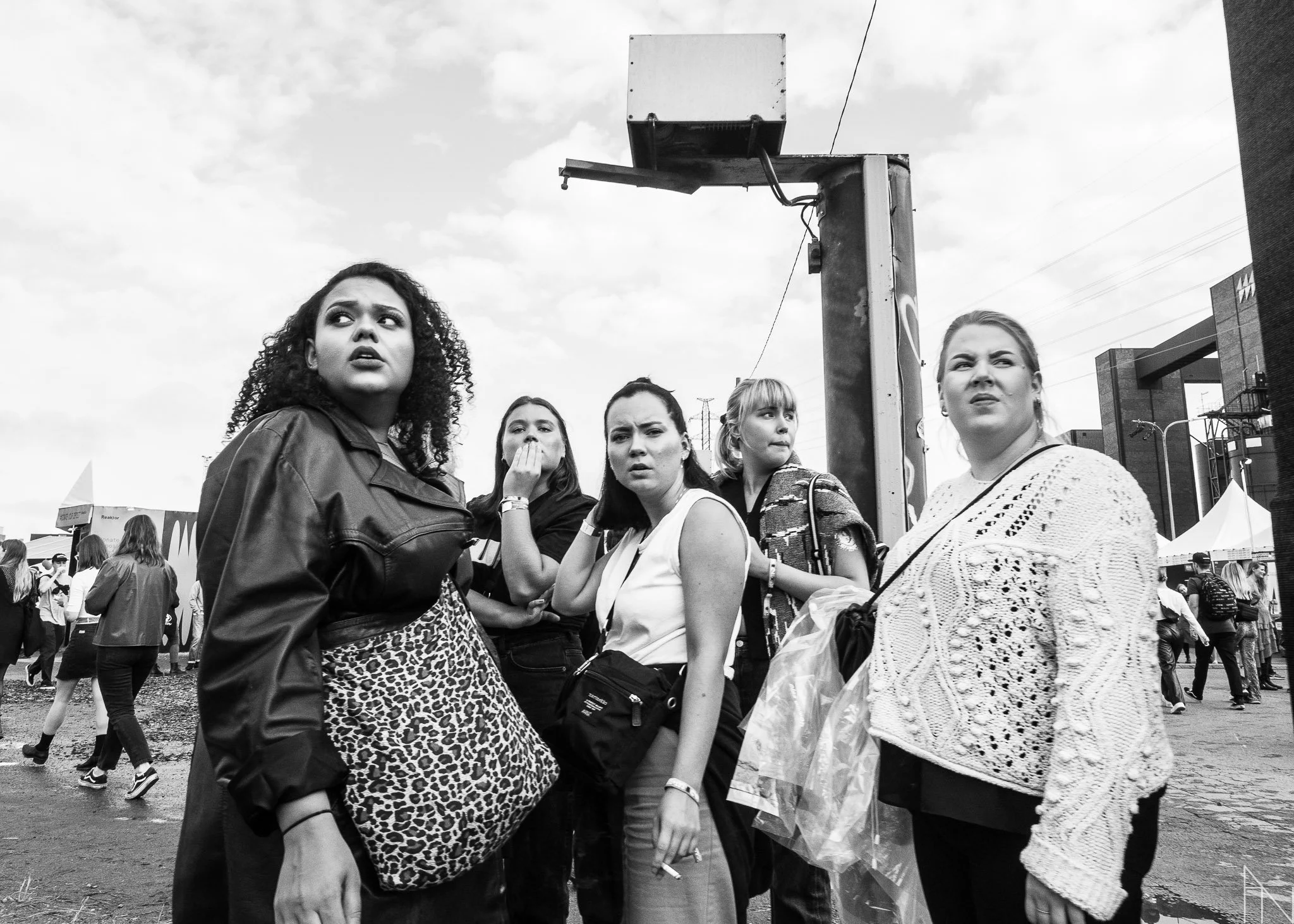 Viisi nuorta naista katsoo jotain epäilevästi tai yllättyneesti. Taustalla muita ihmisiä ja rakennuksia, pilvinen taivas.