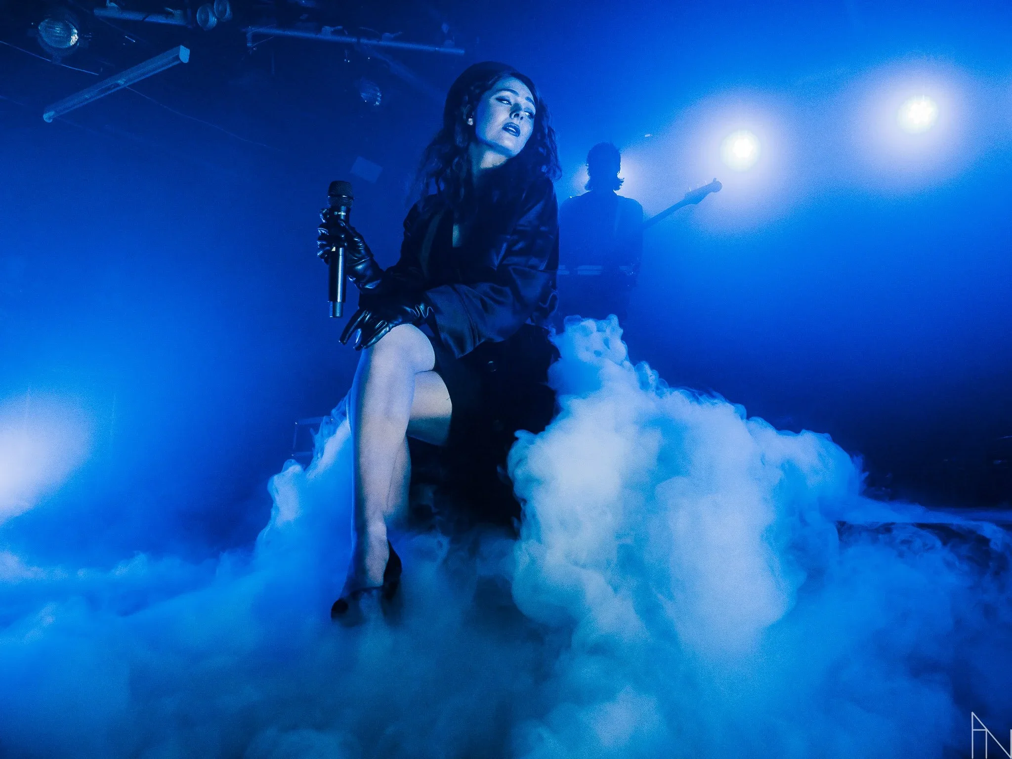 Nainen lavalla, päässä mustat hiuksen, pukeutunut mustaan, istuu korkealla jakkaralla ja pitää mikrofonia kädessään, taustalla kaksi musiikkivirtuoosia ja sininen valaistus. Paljon savua. Behm.