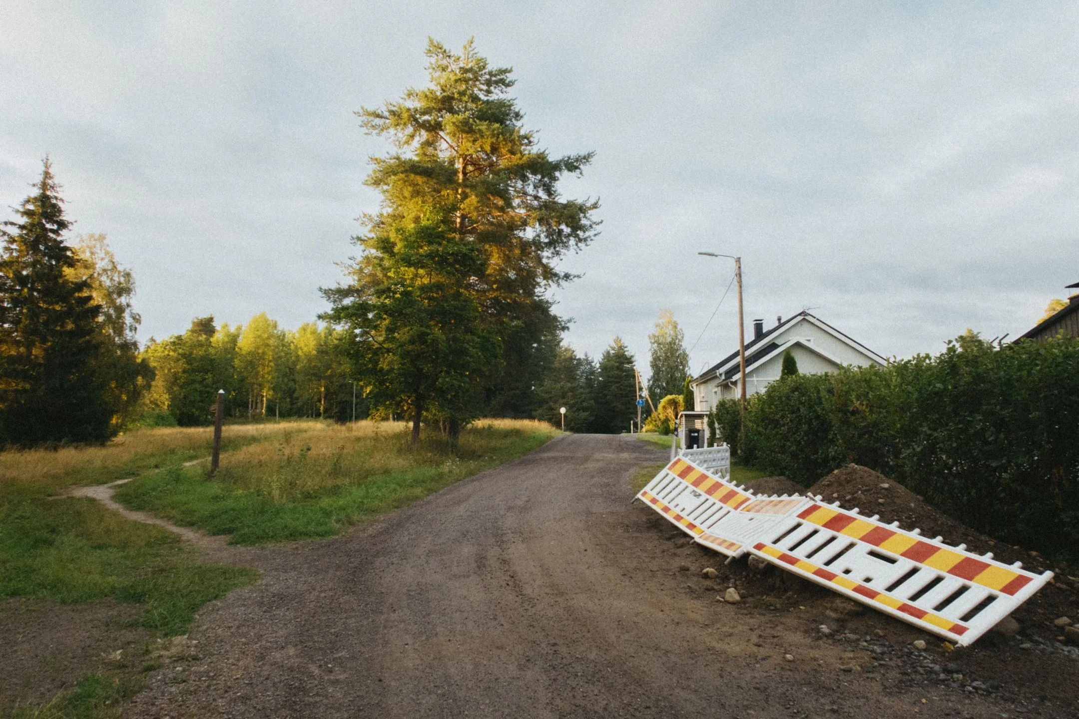 Kuva näyttää pienen asuinalueen hiekkatiepihalle, jossa on rakennustarvikkeita ja ympärillä puita ja taloja.