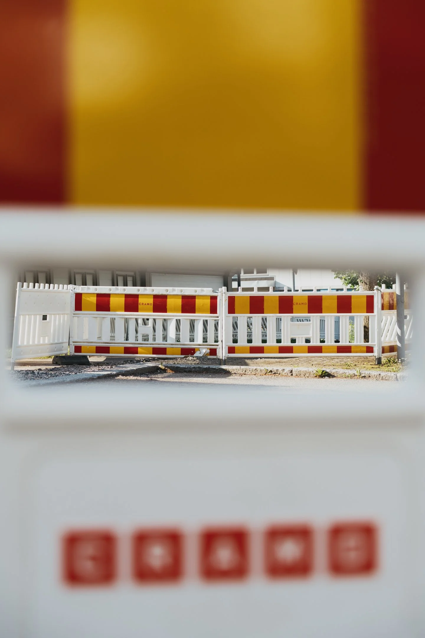 Puutarhakatoksen tai rakennuksen edessä oleva punavalkoinen aita ja pysäköintialue näkyvät kuvasta, kuva on otettu kynnystasolta ja näkymä on rajattu mutta aitaa ja osittain maasta näkyviä kasveja.