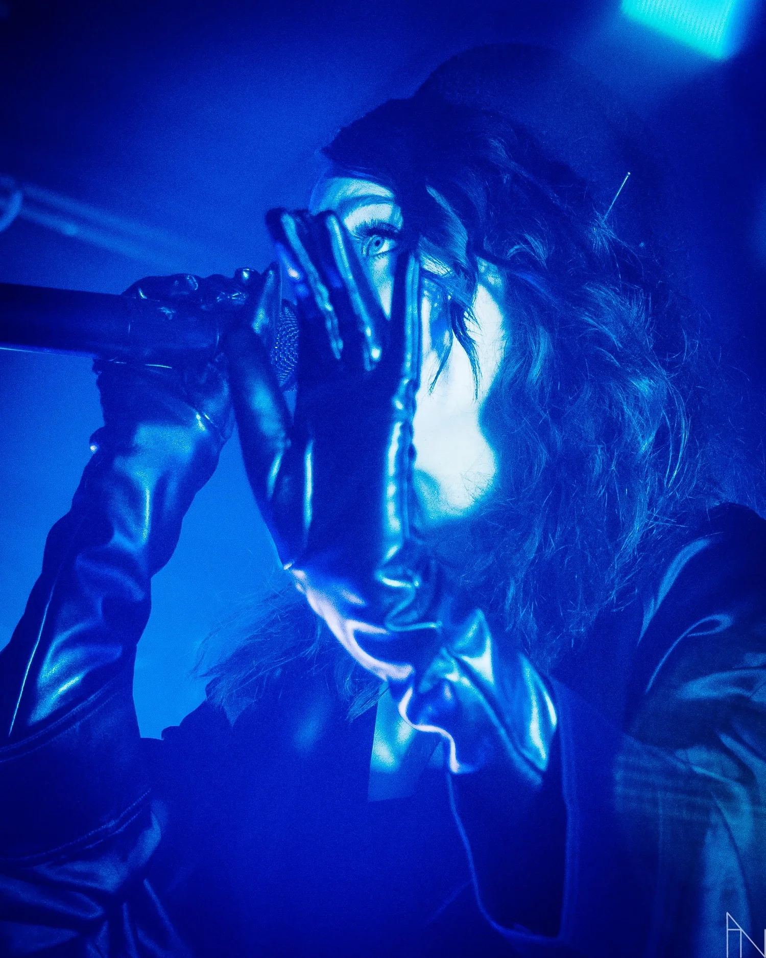 Naista laulaa mikrofoniin rivistetyllä mustalla nahkatakilla ja mustilla käsineillä, valaistuna sinisessä valaistuksessa. Silmä näkyy sormien välistä.