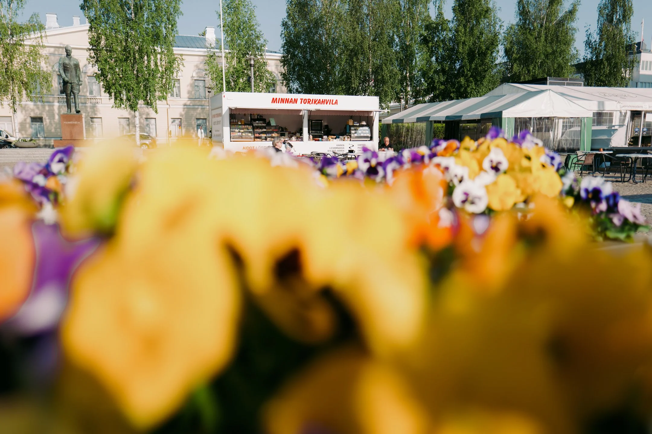 Kuva etualalta, jossa on kukkakimppuja ja markettiosasto taustalla. Taustalla näkyy kauppakioskia ja taulut, jotka mainostavat Minnan Torikahvila. Taustalla on puutarha ja rakennuksia. Mikkelin tori.