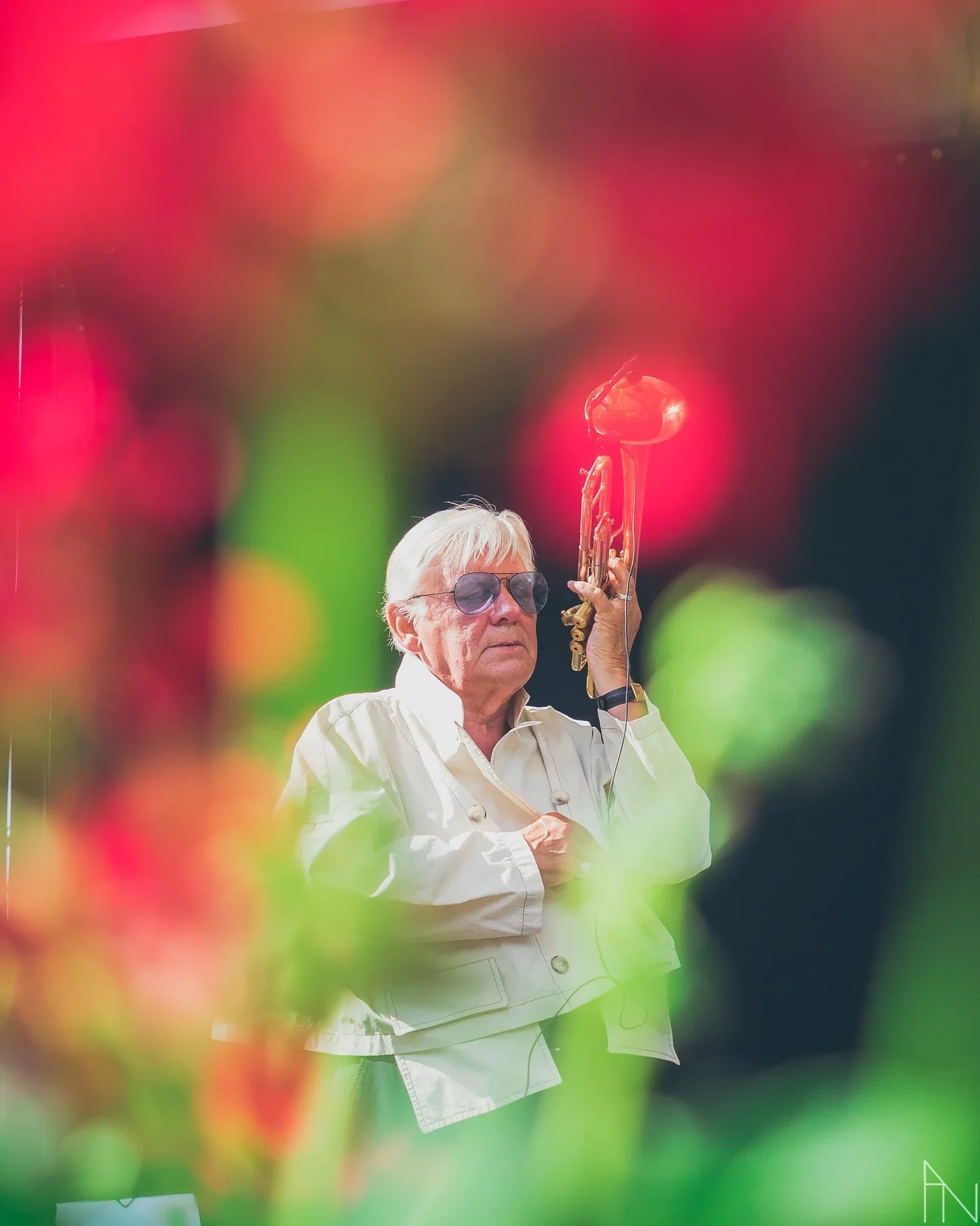 Ikoninen mies soittaa saksofonia, pukeutuneena valkoiseen takkiin ja tummiin laseihin, etualalla värikkäitä kukkia ja epätarkka tausta. Pori Jazz.