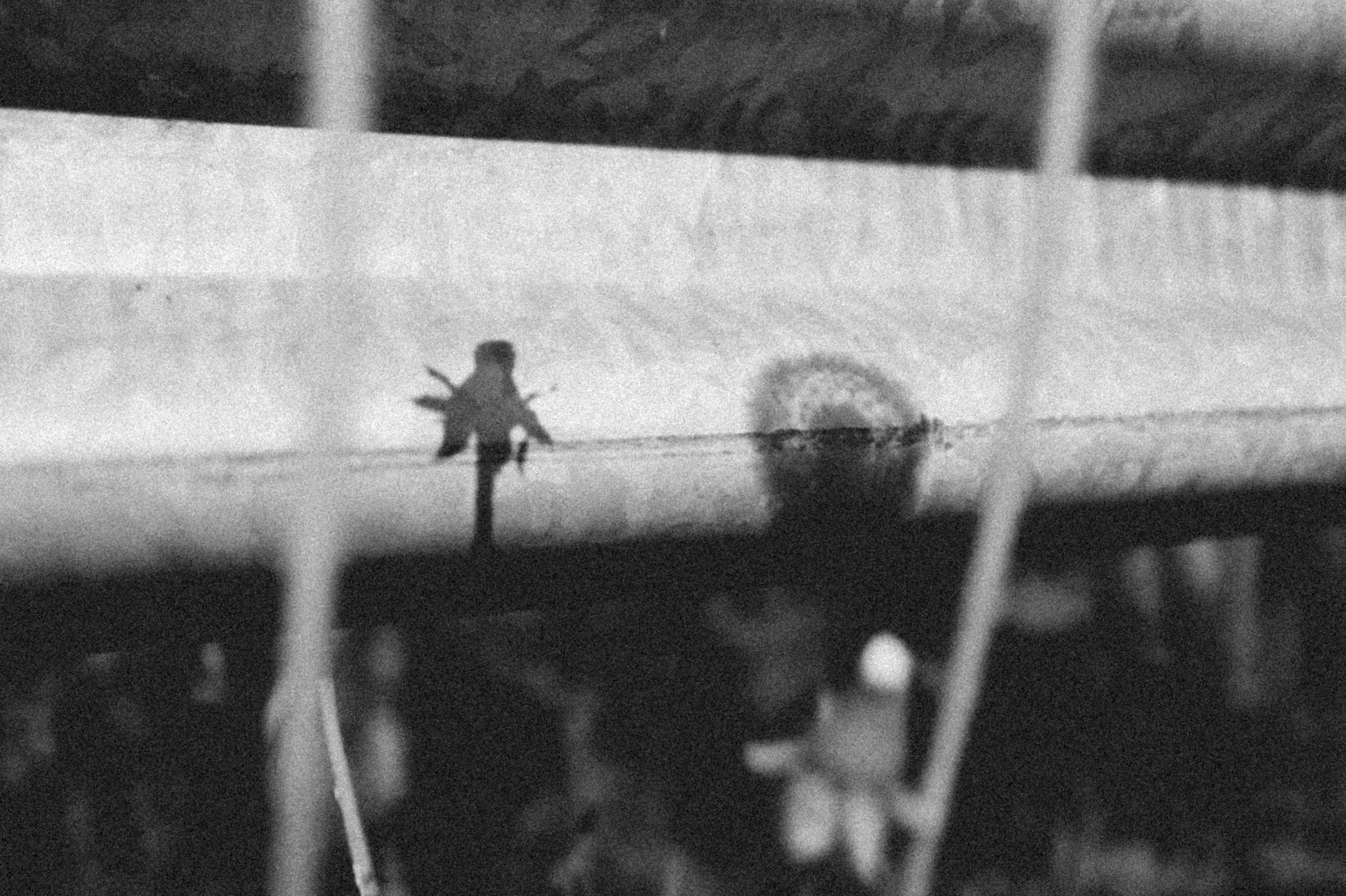 Mustavalkoinen kuva, jossa näkyy pienoismaalaisnäkymä usealla tasolla. Korkealla on pieni mieshahmo, joka kävelee tai seisoo, ja alhaalla on muita ihmishahmoja ja rakennuksia, todennäköisesti kaupunginäkymä.