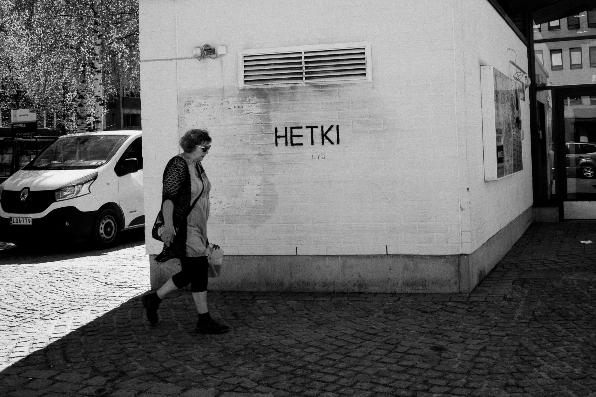 Nainen kävelemässä kadulla, taustalla auto ja rakennus, jossa on graffiti, jossa lukee 'HETKI'.