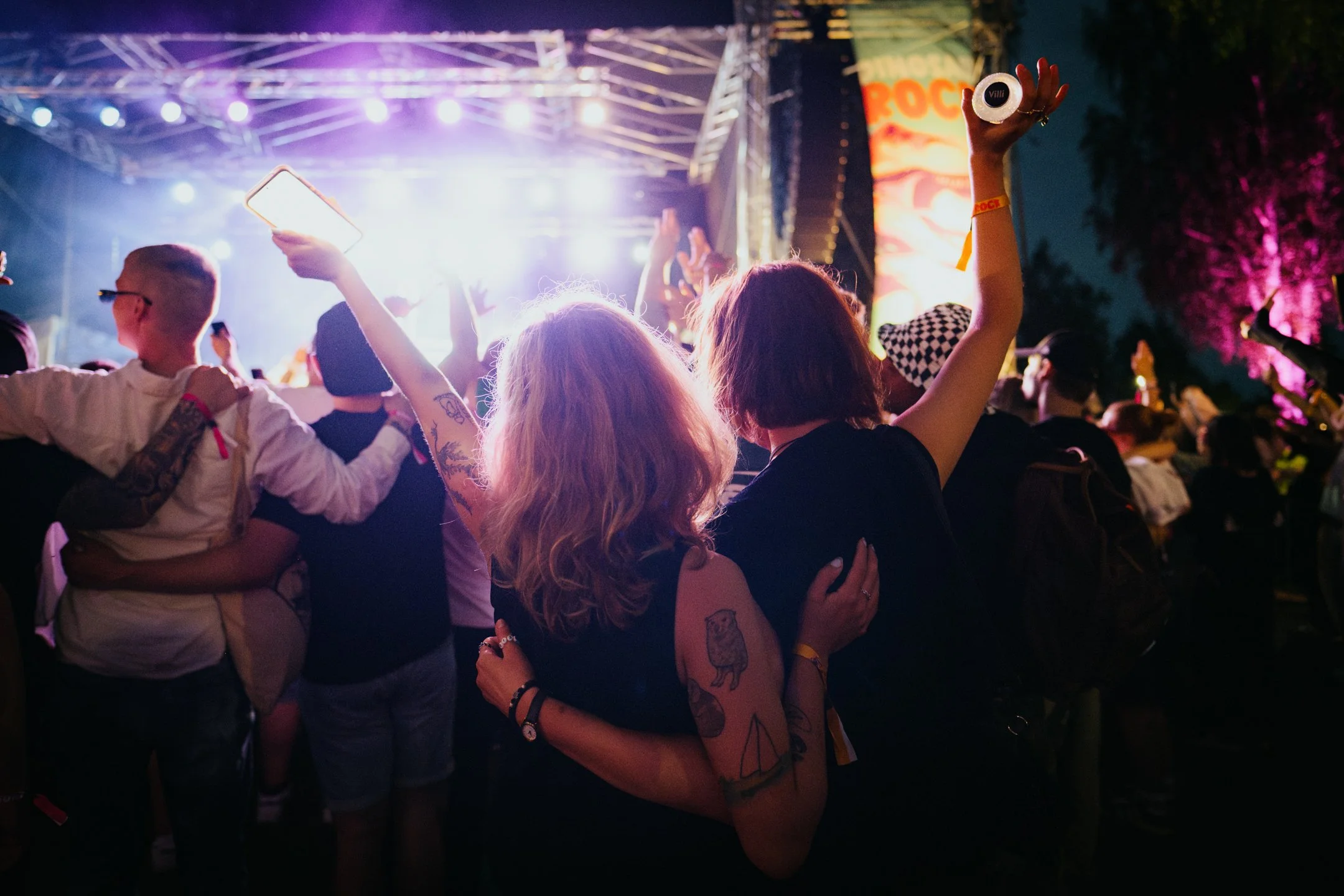 Ihmiset nauttivat musiikkifestivaalista illalla, tanssivat ja ottavat selfieitä lavan edessä, värikkäitä valoja ja tummia puita taustalla.