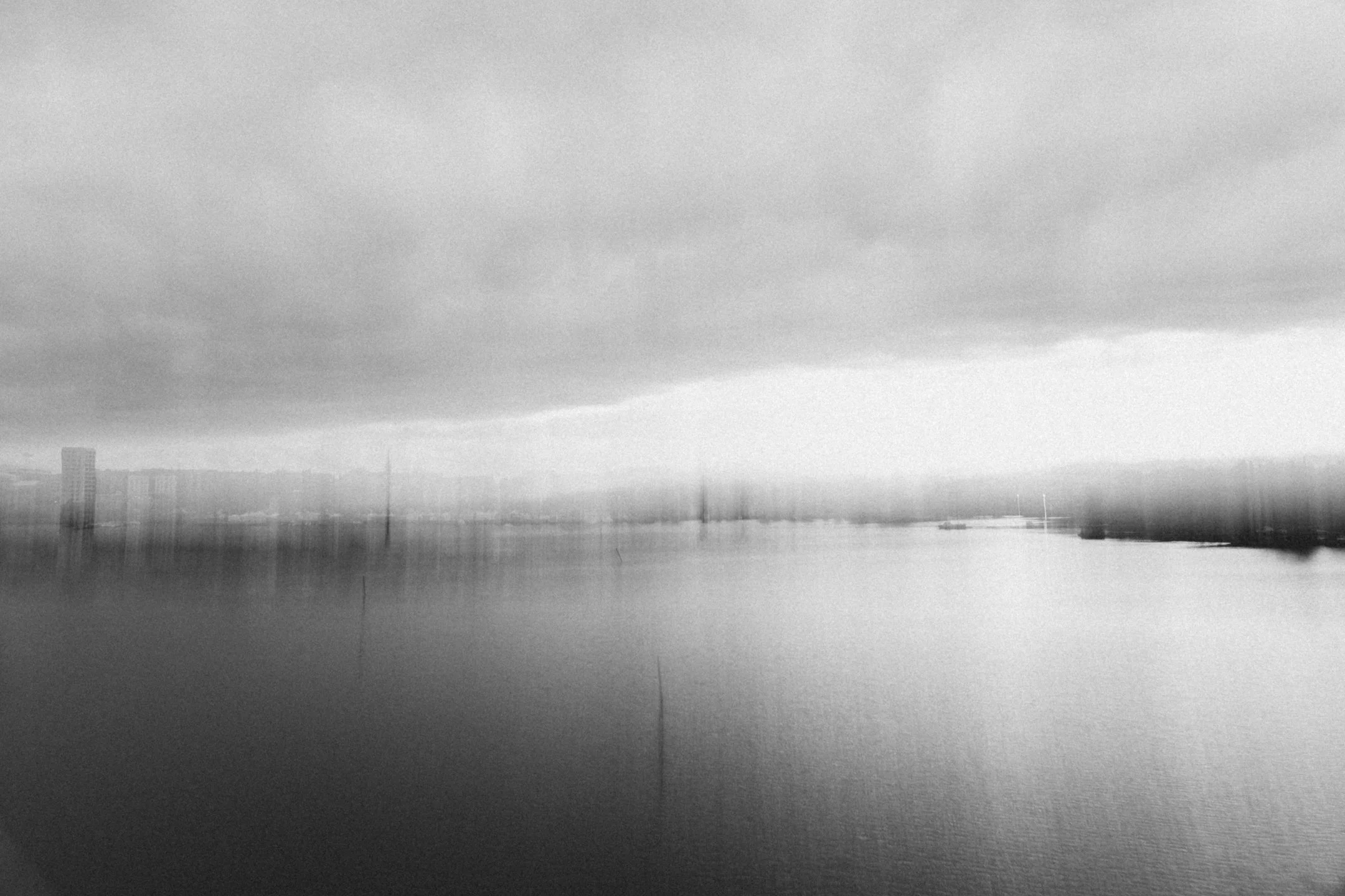 Mustavalkoinen kuva järvest ja kaupunkimaisemasta taivaan alalaidassa, pilvet harmaat ja tiiviit, veden pinnalla heijastuksia rakennuksista ja laivoista.