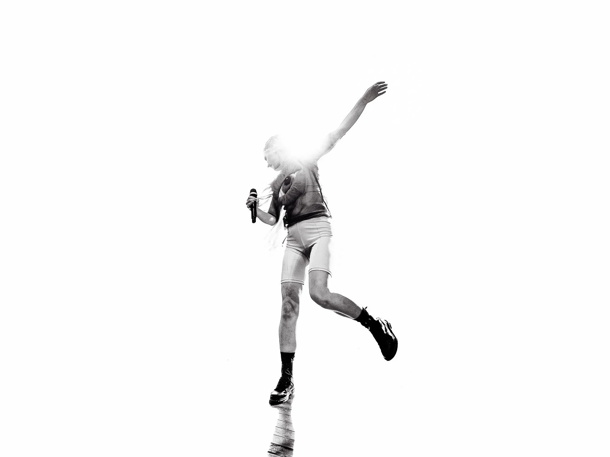 Mustavalkoinen kuva vapaana olevasta ihmisestä, joka tanssii tai esiintyy, pitäen mikrofonia, taustalla valkoinen tyhjä tila.