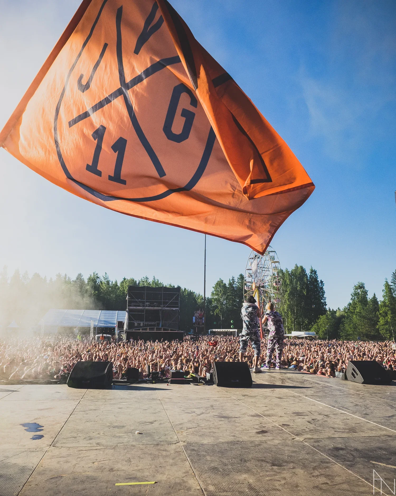 Festivaalialue, jossa suuri onnellinen yleisö ja lavalla kaksi esiintyjää. Taustalla sirkusteltta ja laiva-raidepyörä. Etualalla suuri JVG-lippu.