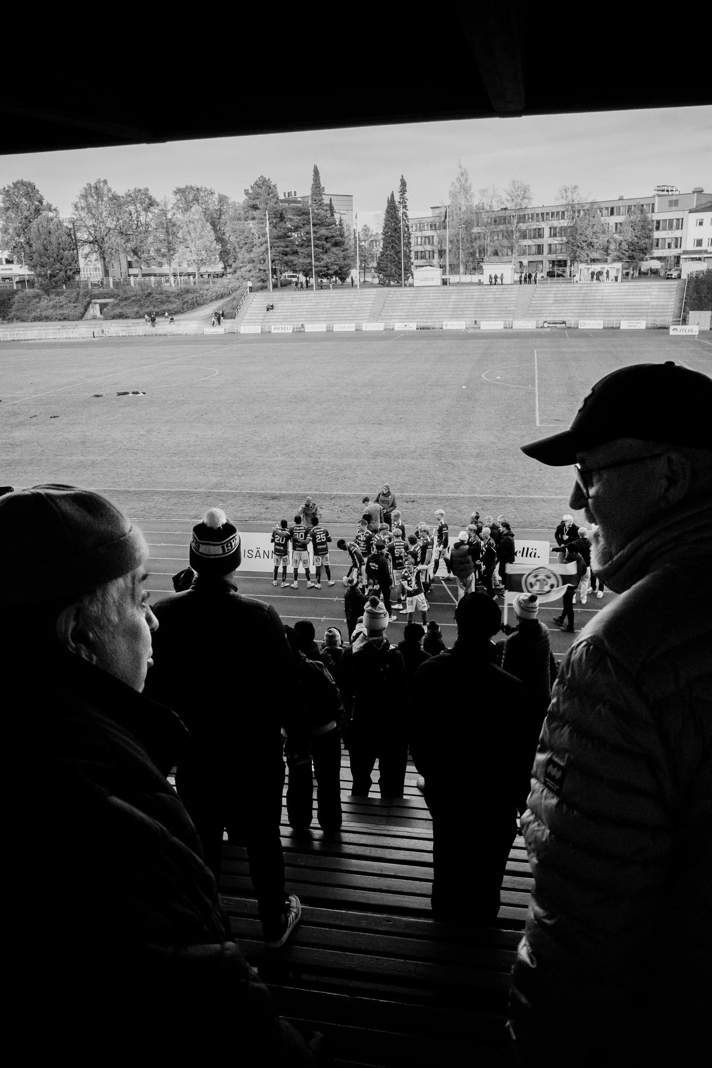 Mustavalkoinen kuva katsomosta jalkapallostadionilla, jossa katsojat seuraavat peliä ja pelaajat keskellä aluetta