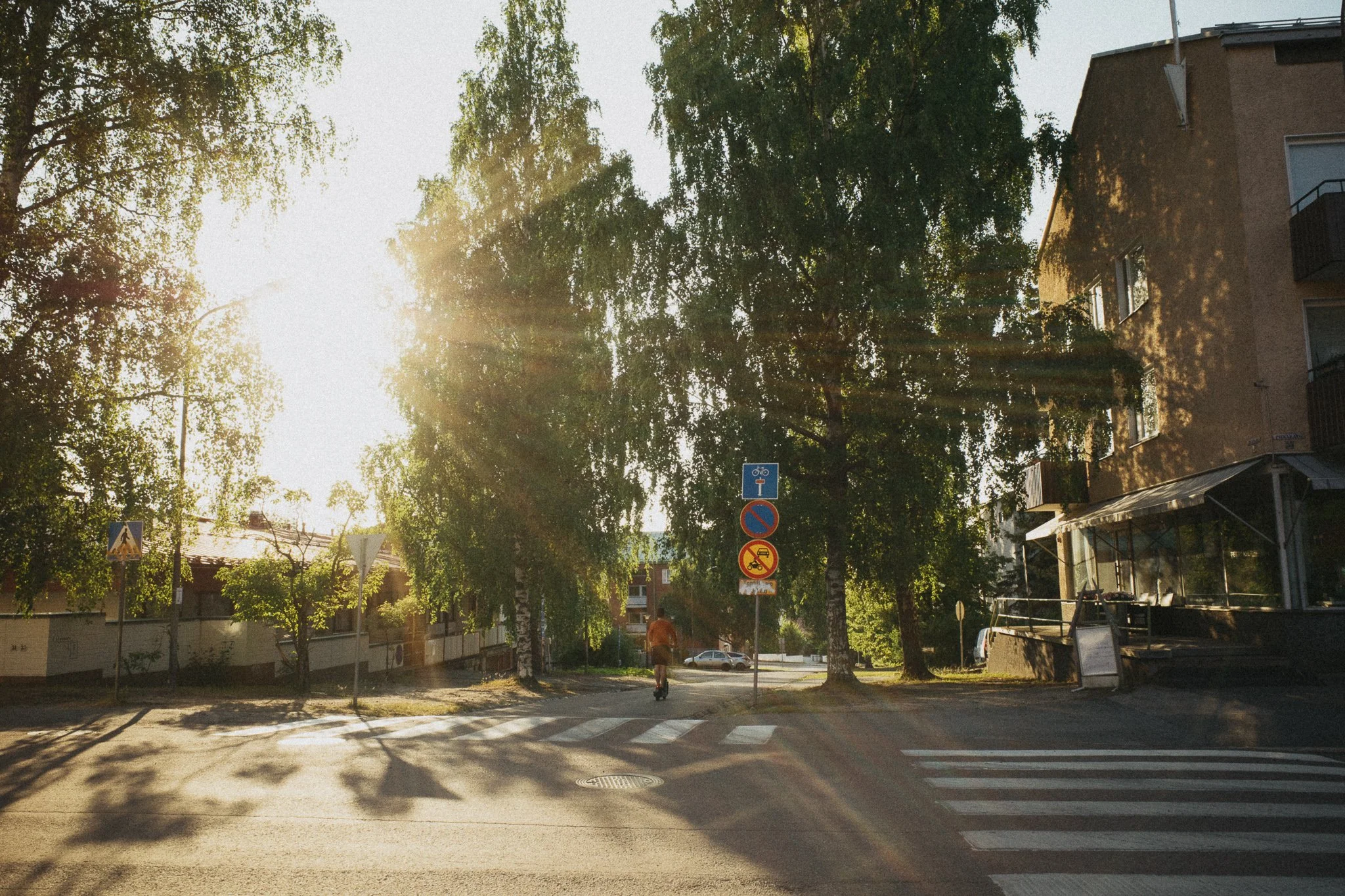 Kuvassa kaupunkialueen katu ja risteys auringonlaskun aikaan. Näkyvissä on puita, jalankulkijansillan ja liikennemerkkejä. Jyväskylä.