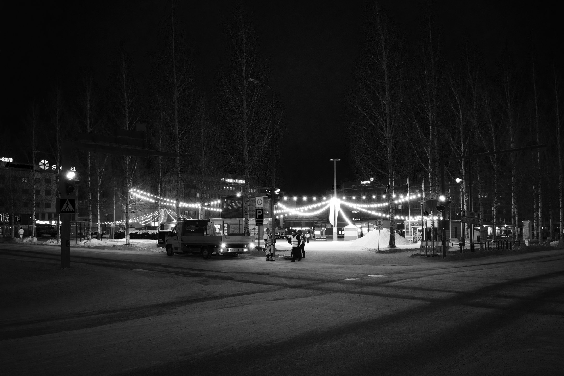 Yökuva tapahtumalasta, jossa on jouluvaloja, ihmisiä ja ajoneuvoja, lumista maata ja puita, kauppojen valoja taustalla.