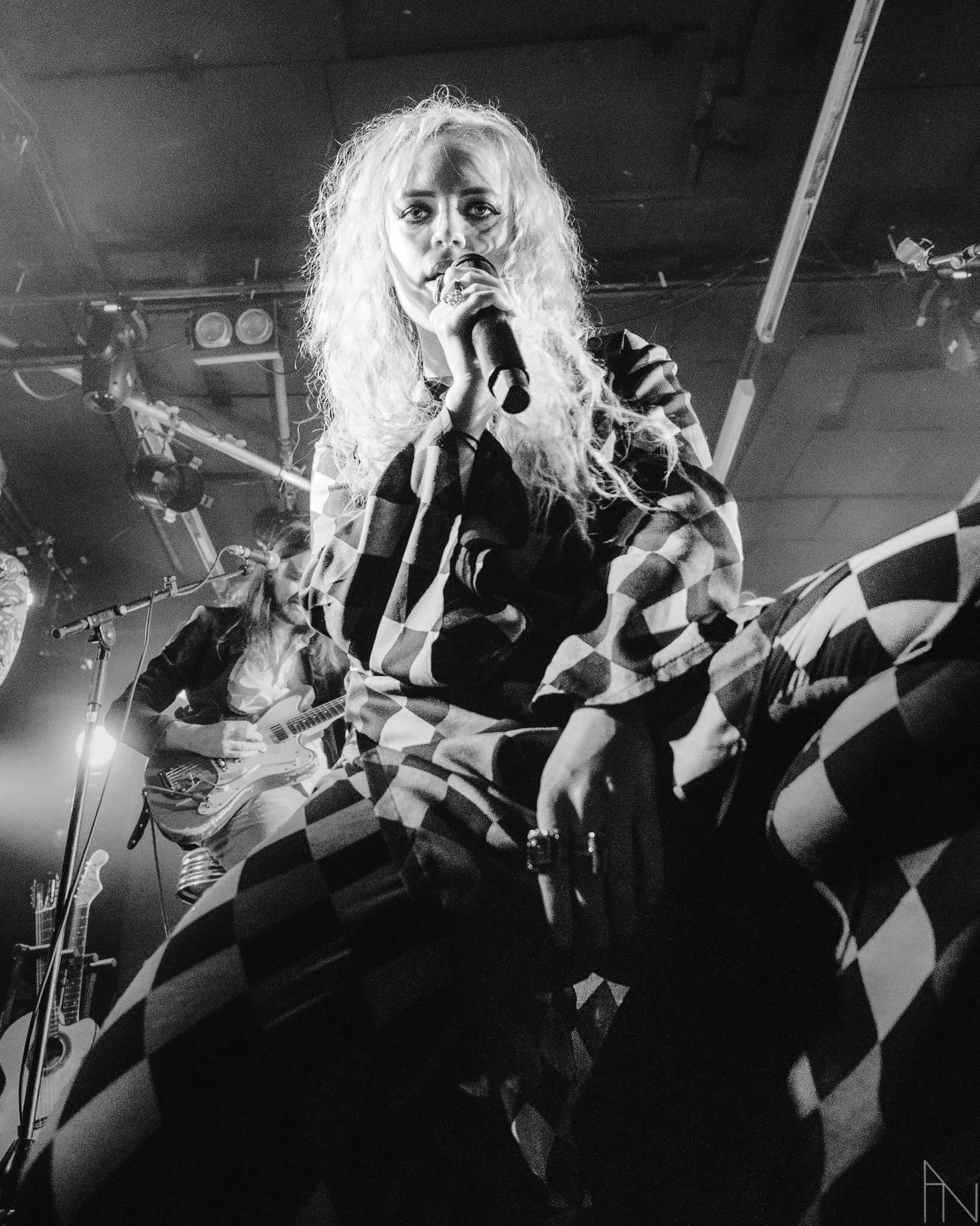 Elli Haloo katsoo suoraan kameraan, toinen jäsen soittaa kitaraa, ympärillä valoja ja musiikkilaitteita, mustavalkokuva.