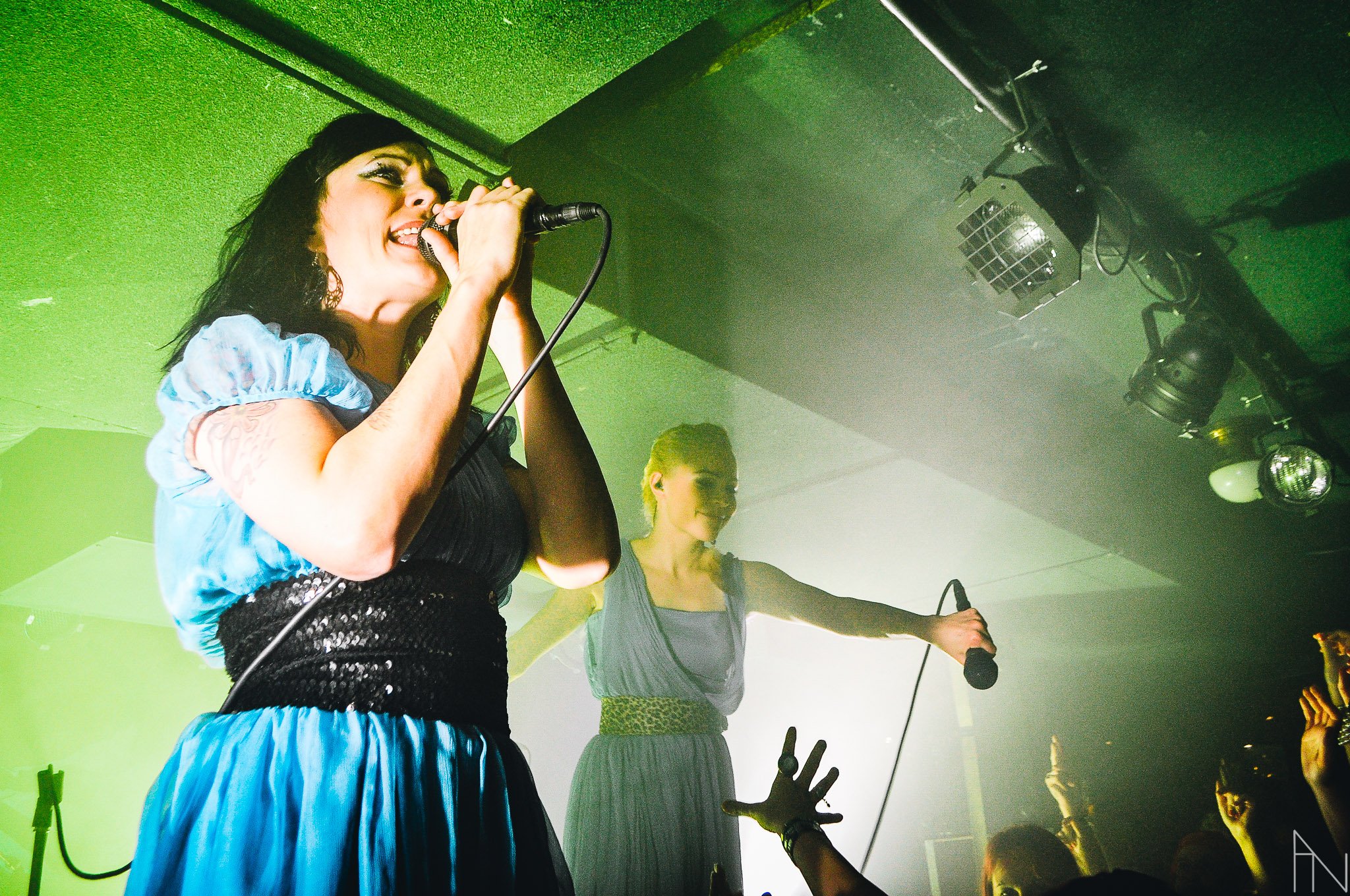 Kaksi naista laulamassa mikrofonit hyllyssä klubilla tai keikalla. Yleisöä, joilla on käsiä ylhäällä, ja valoefektejä taustalla. PMMP Mikkeli 2009.