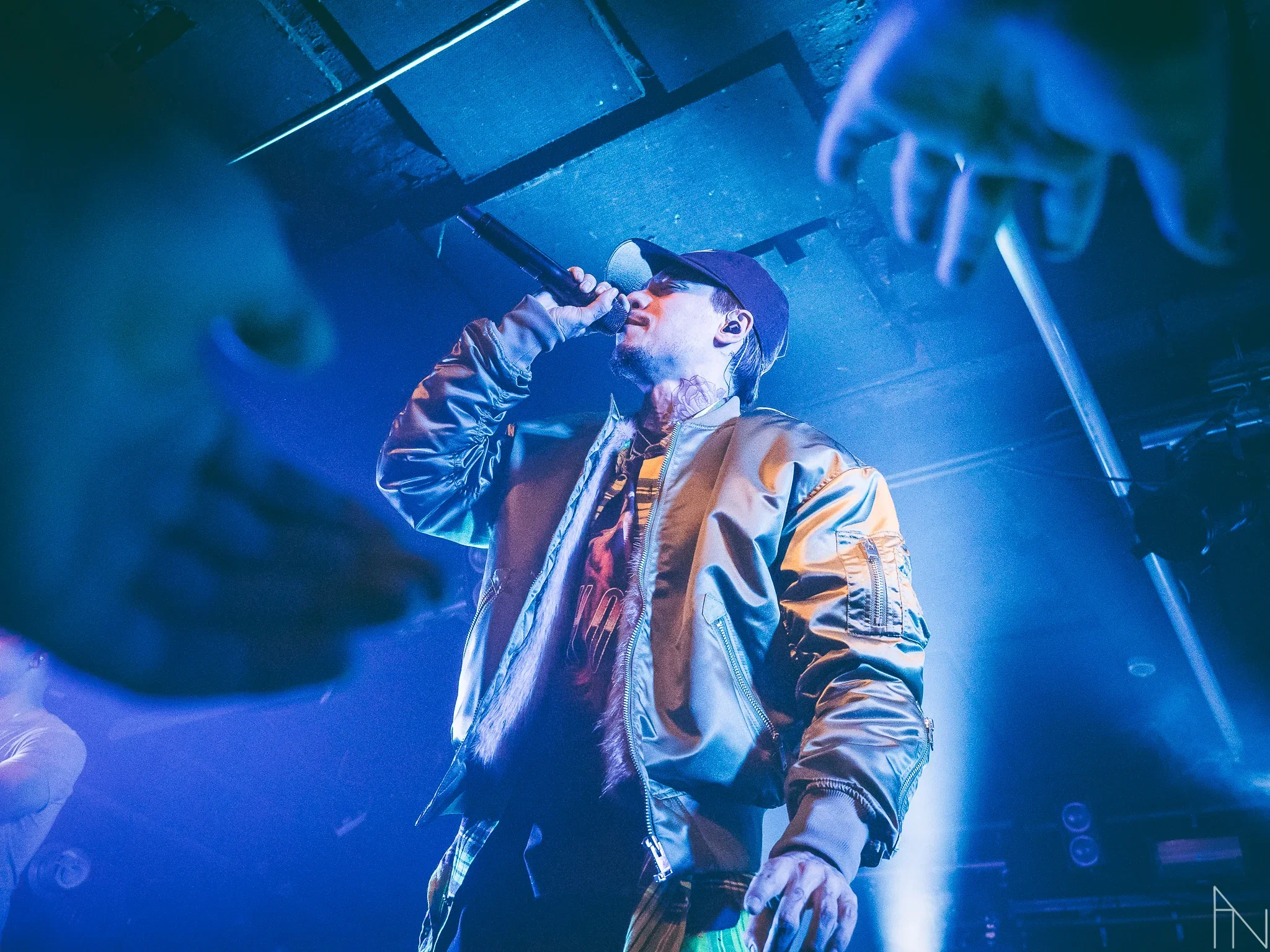 Artisti esiintyy lavalla, käyttäen lakkia ja nahkatakkia, laulaen mikrofoniin valaistuksen alla. Kuvan ulkopuolelta tulevat kädet koittavat koskettaa esiintyjää. Mikael Gabriel Lutakko.