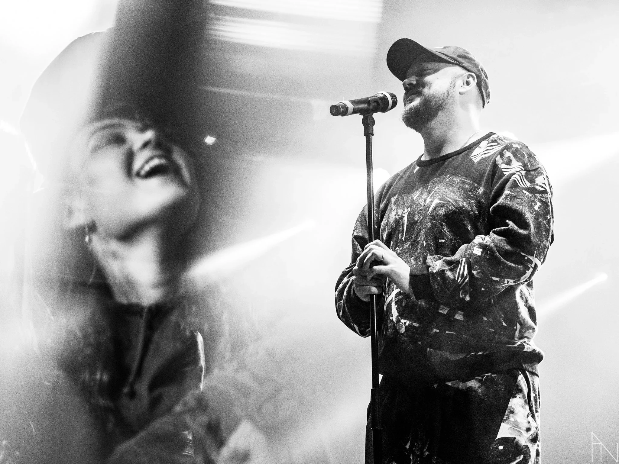 Mies laulaa mikrofonin äärellä, nainen hymyilevän naaman ikonin lähellä. Mustavalkokuva.