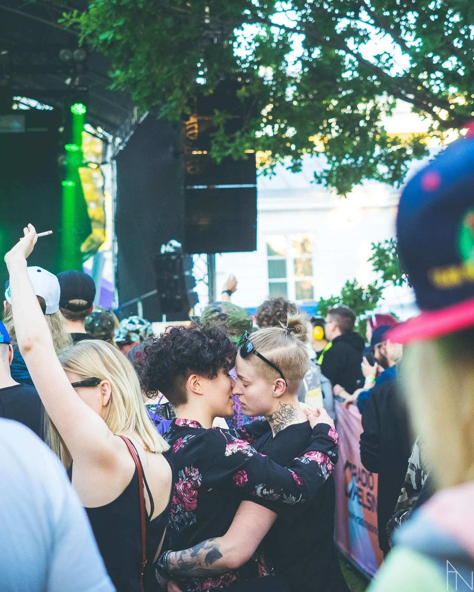 Parsing crowd at an outdoor music concert, with two women embracing and about to kiss in the foreground, surrounded by dancing and enjoying attendees. Ihmisten edessä.