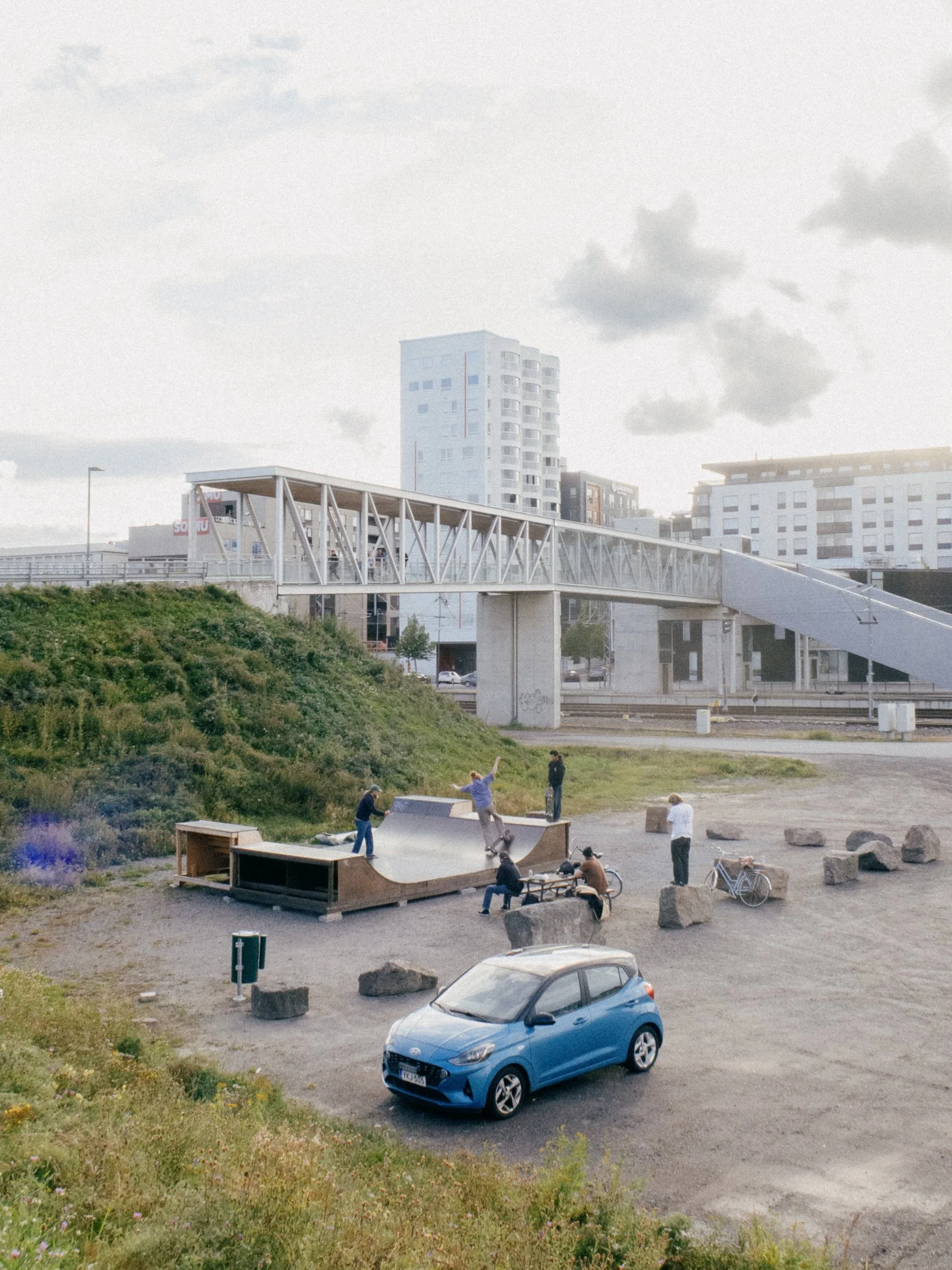 Kuva kaupungin ulkopuolisesta parkkipaikasta, jossa on skateboard-rampteja, ihmisiä jotka skeittaavat ja istuvat, ja autot sekä pyörät.