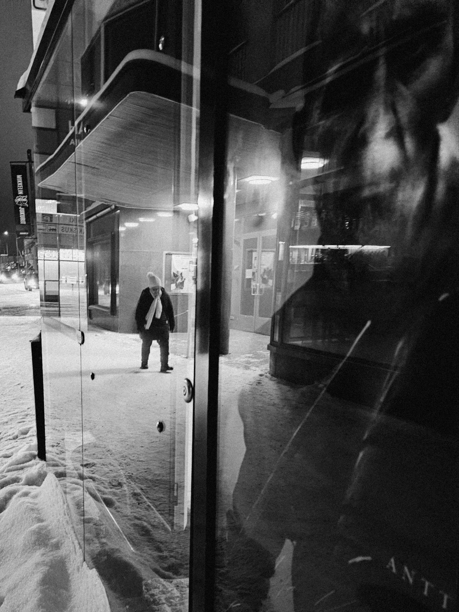 Mustavalkoinen kuva kaupunkipysäkillä lumen ja jään keskellä. Näkyy keski-ikäinen nainen käyttäen huivia ja takkia odottamassa pysäkillä. Näkyy myös toinen osittain näkyvä henkilö heijastuksessa ikkunassa.