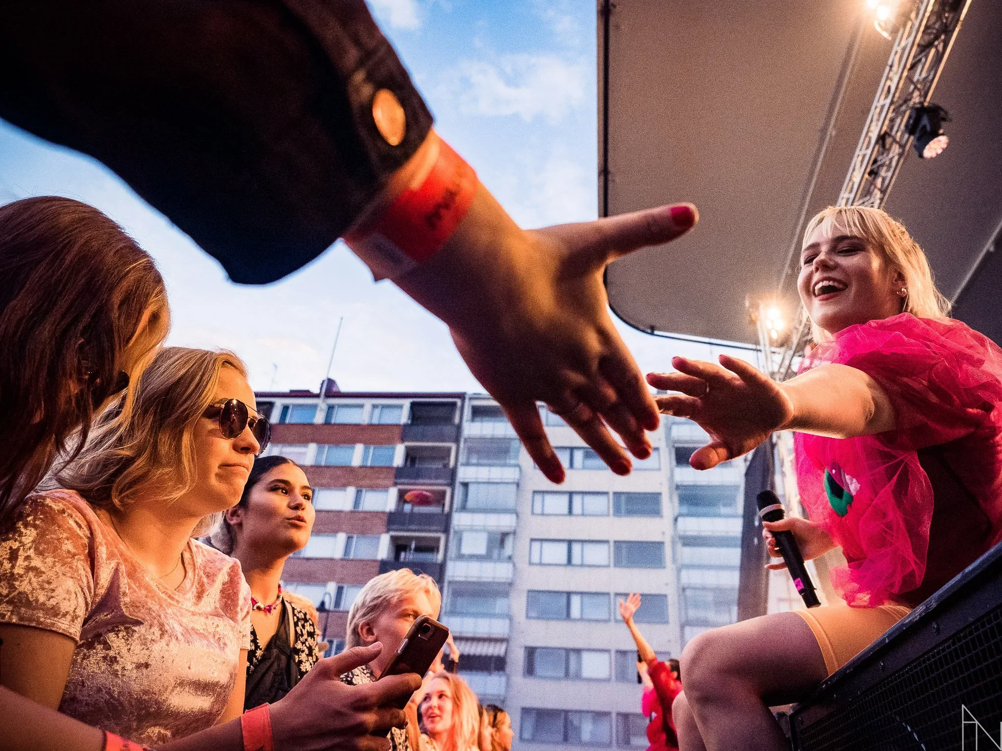 Nuori nainen laulaja kyselee yleisöstä ja antaa kätensä yleisölle. Taustalla useita ihmisiä ja kerrostaloja, tapahtuma ja valoisuus ulkoilmassa.