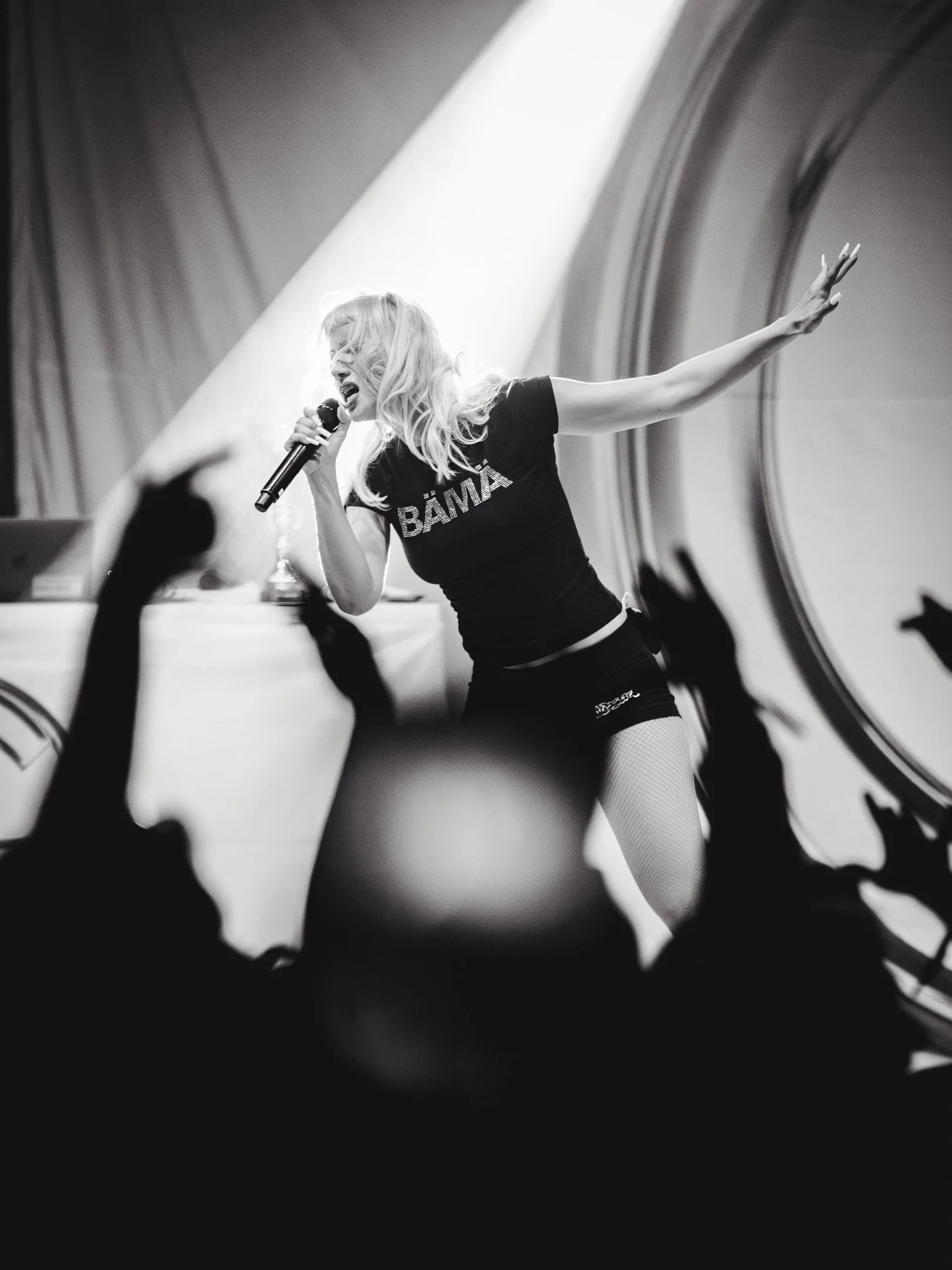 Nainen esiintymässä lavalla laulamalla mikrofoni kädessä, fanit kohottavat käsiään etualalla, mustavalkoinen kuva. Olga Dinosaurock 2025 Mikkeli.