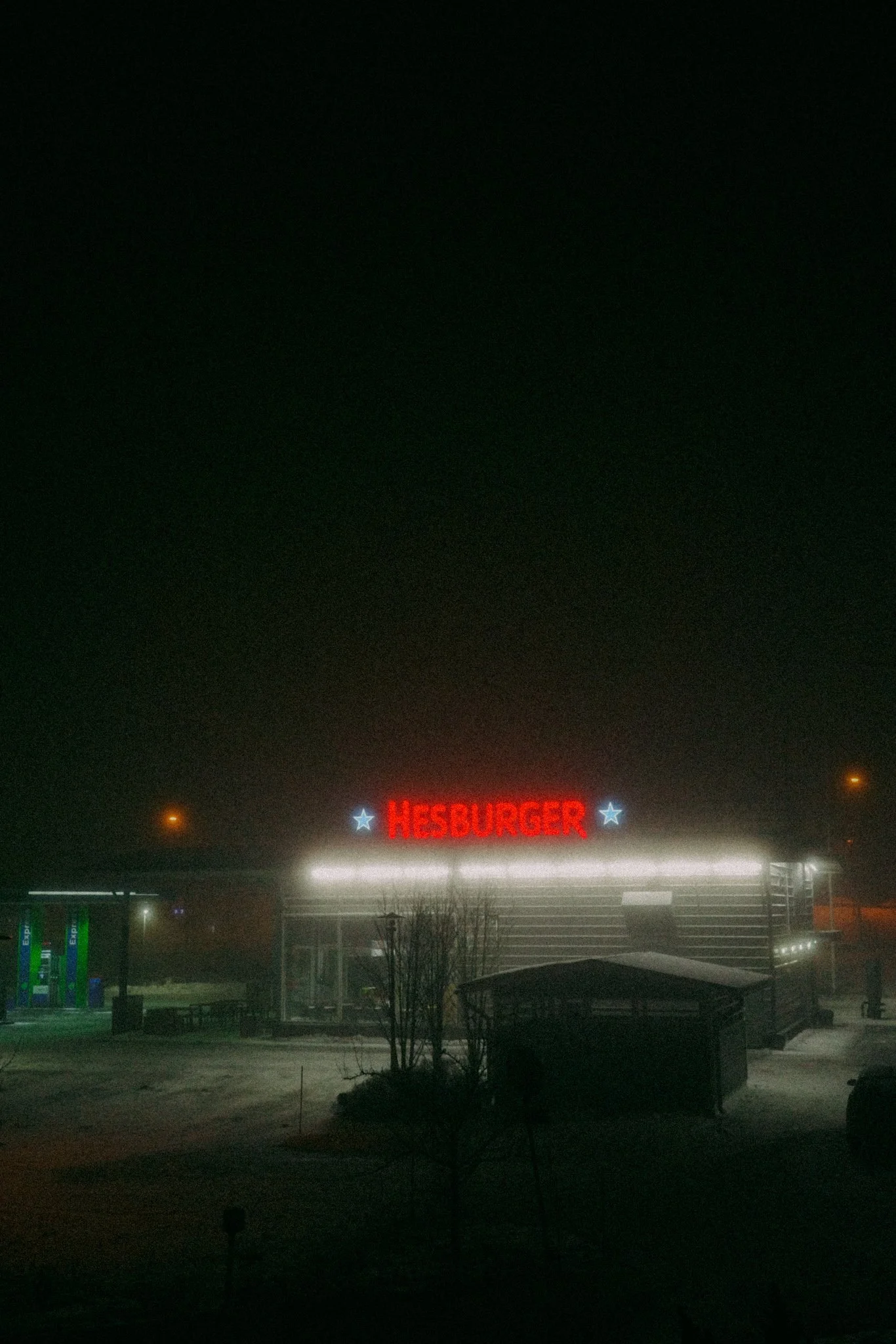 Yöllinen kuva Hesburger-ravintolan kyltistä, jossa näkyy punainen nimi ja kaksi sinistä tähteä. Osa pumppausasemasta ja puita edessä.