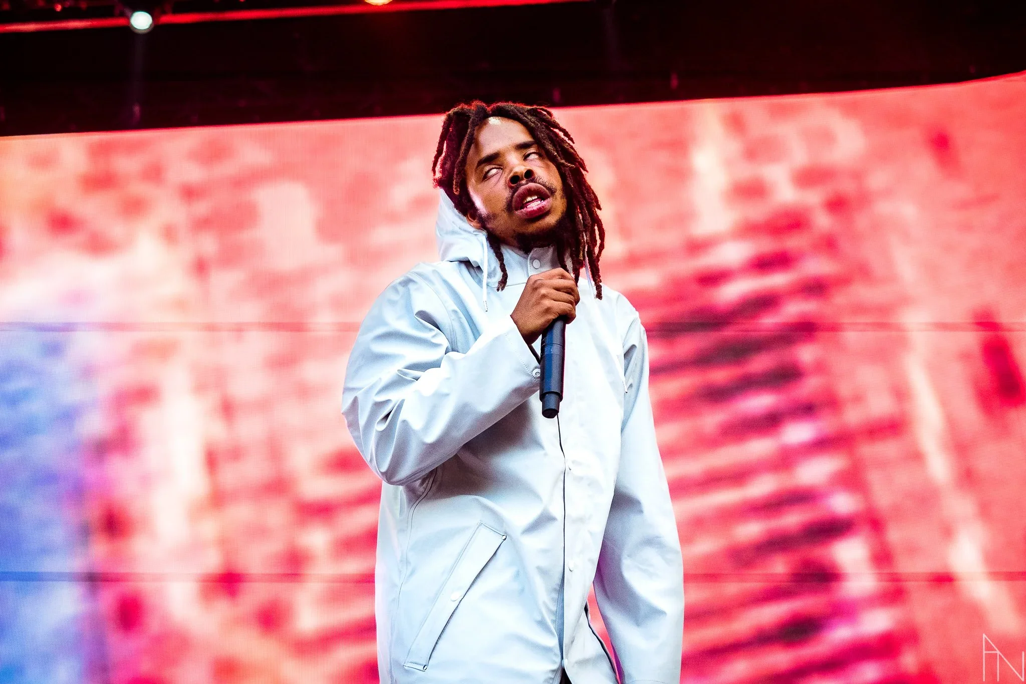 Mies esiintyy lavalla, pitää mikkiä ja näyttää yllastyyneeltä, taustalla suuri punainen näyttö tai valo.