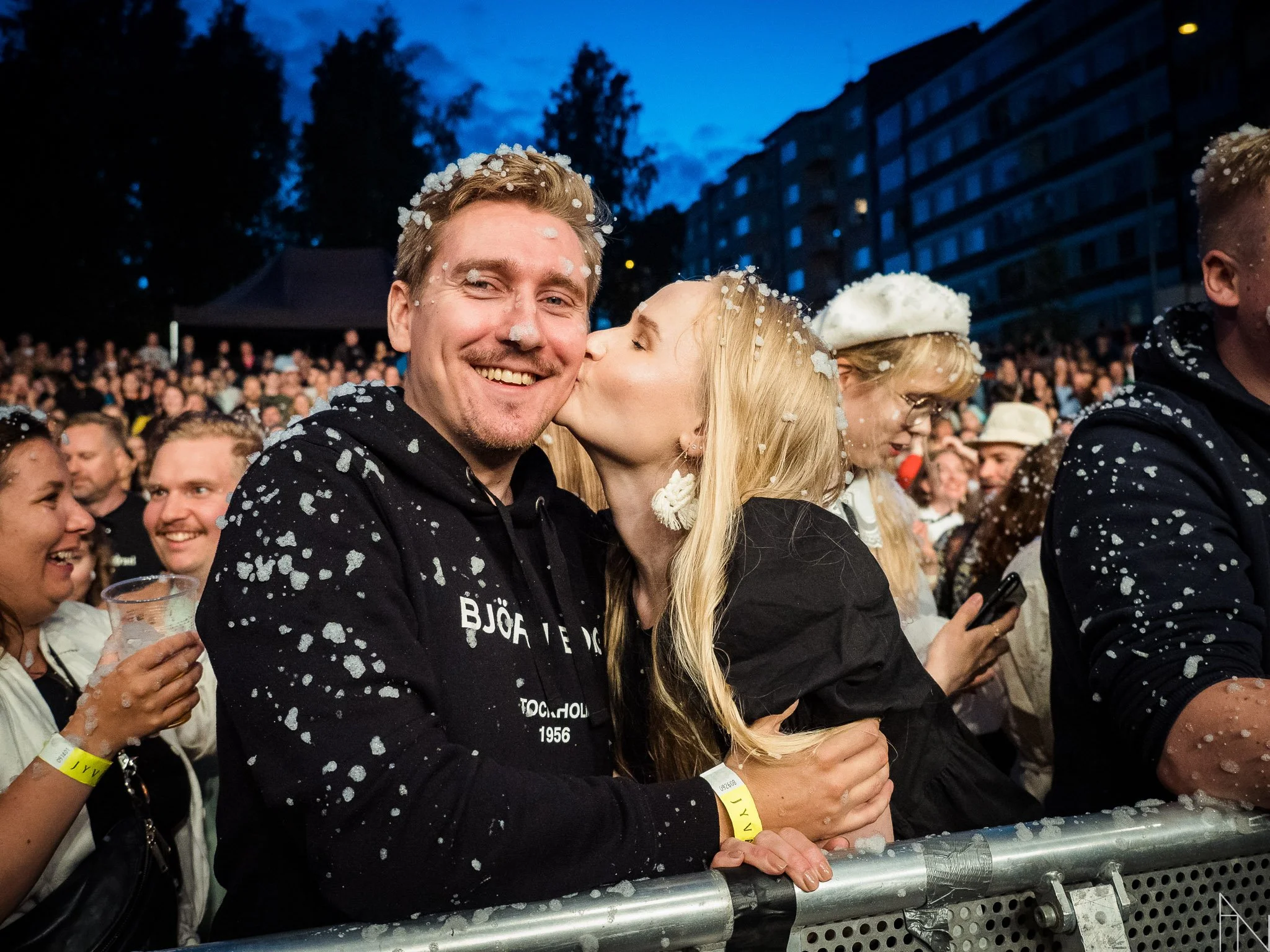 Ihmiset nauttivat festivaalitunnelmasta, yksi nainen suukottaa miehen poskea, jossa on lavassa ja ihmisiä taustalla. Puistojuhlat Jyväskylän Kesä.