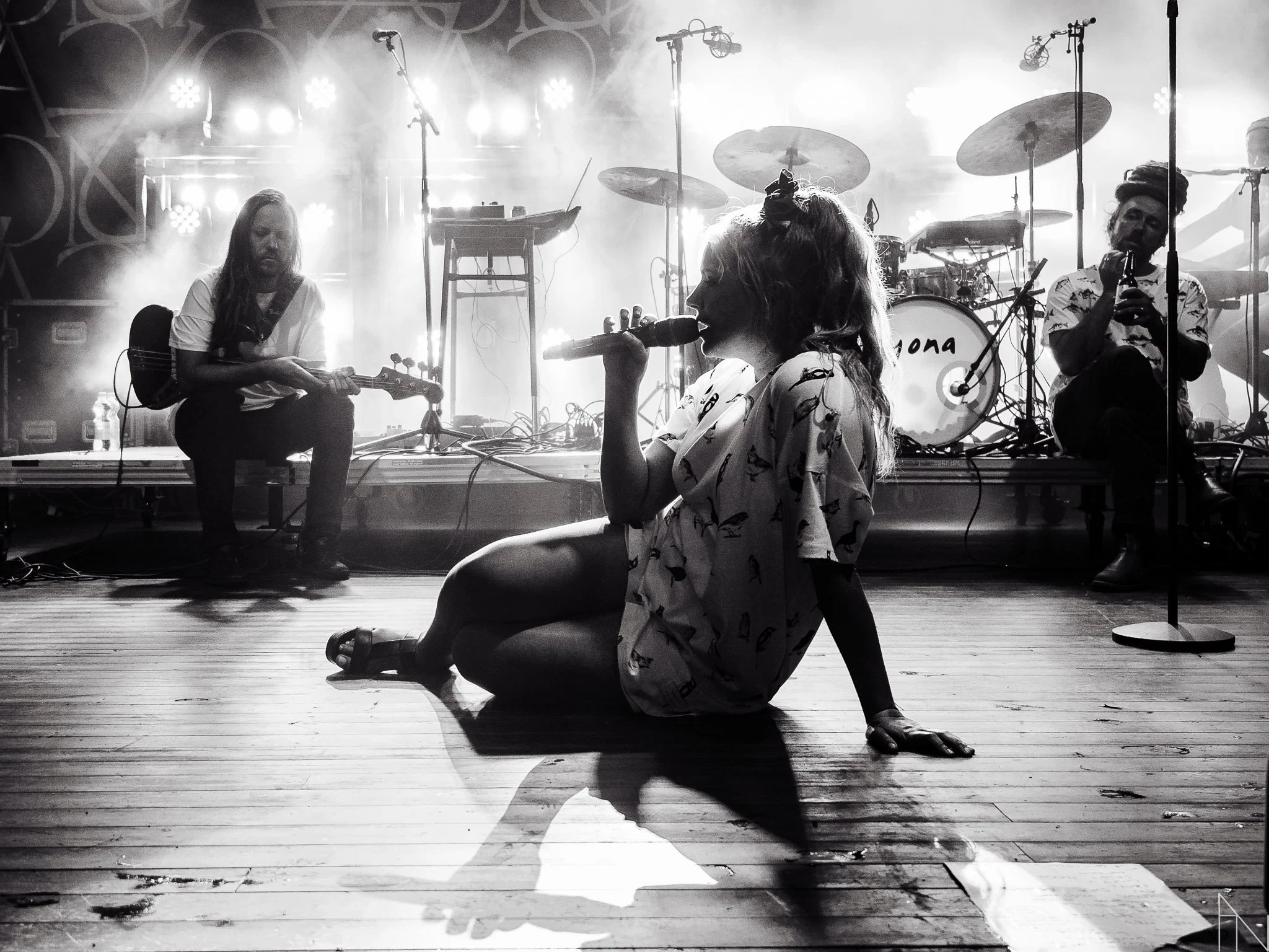 Naisharrastajia esiintymislavalla, yksi istuu lattialla laulamalla mikrofoniin ja toiset soittavat instrumentteja taustalla, valoefektit.