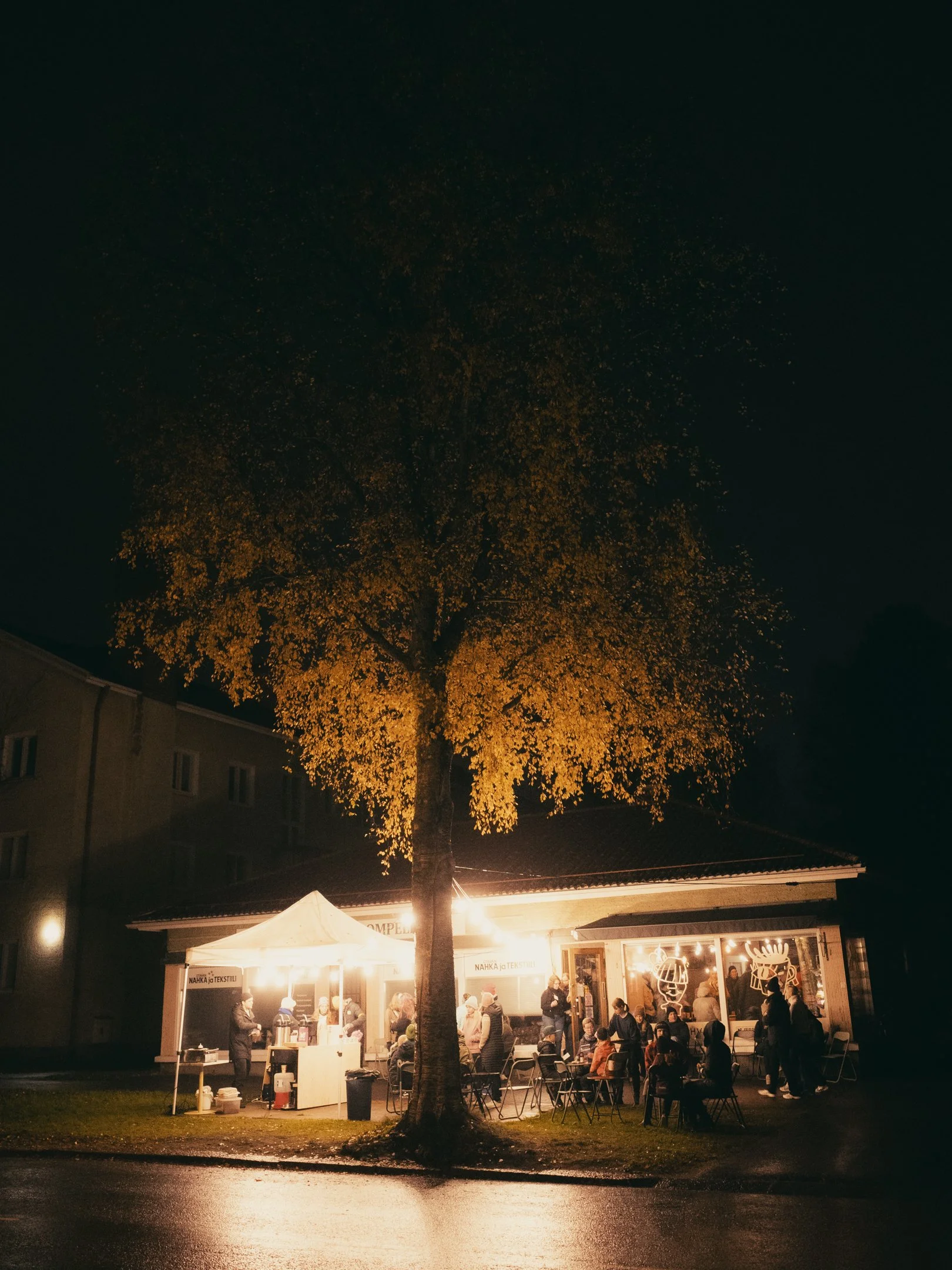 Yökuva esiintymisestä ulkona, jossa on suuri puu ja rakennus, jonka ikkunoista näkyy sisävalot. Paikalla on ihmisiä istumassa ja seisomassa, sekä kojuja ja valoja.