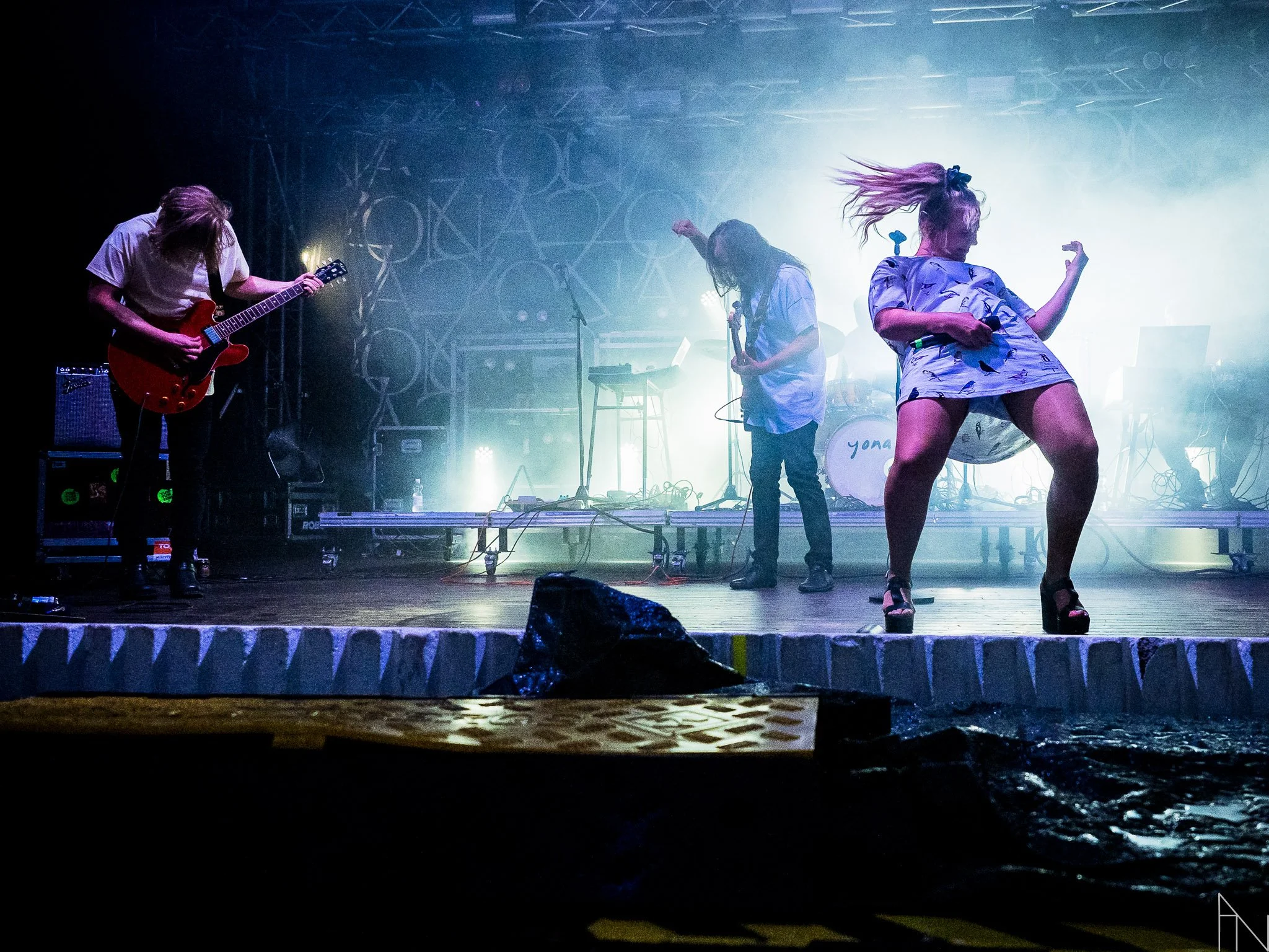 Live rock- ja pop-musiikkikonsertin esiintyjät lavalla, yksi naislaulaja tanssii energisesti, kaksi miehenä laulaa ja soittaa kitaraa taustalla, lavan taustalla valo ja tekninen laitteisto.