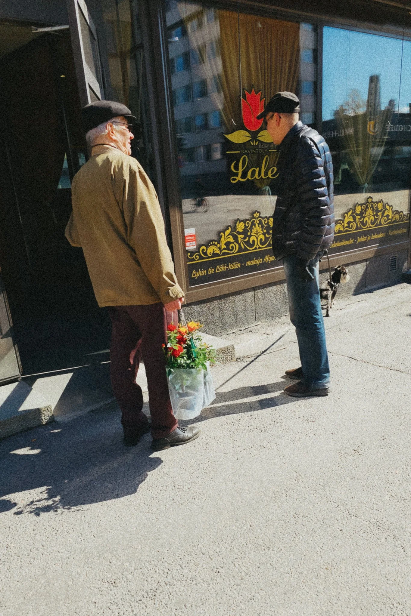 Kaksi miestä keskustelemassa kadulla, toisen miestä kantaa kukkakimppu, taustalla ravintolan ikkuna ja pieni koira.