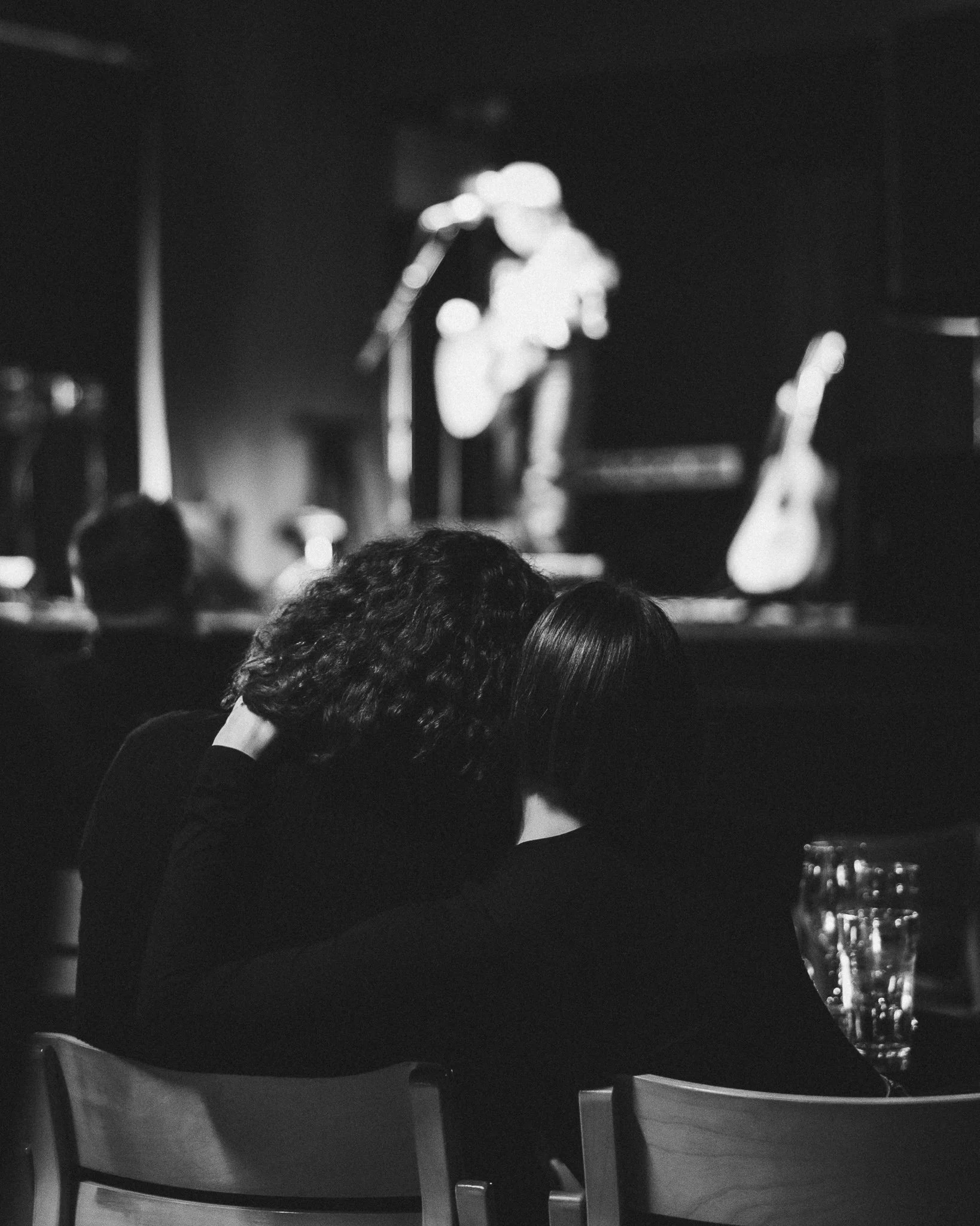 Mustavalkokuva nuoresta parista, jotka istuvat yhdessä katsomassa konserttia, taustalla kitara- ja mikrofonilaitteet.