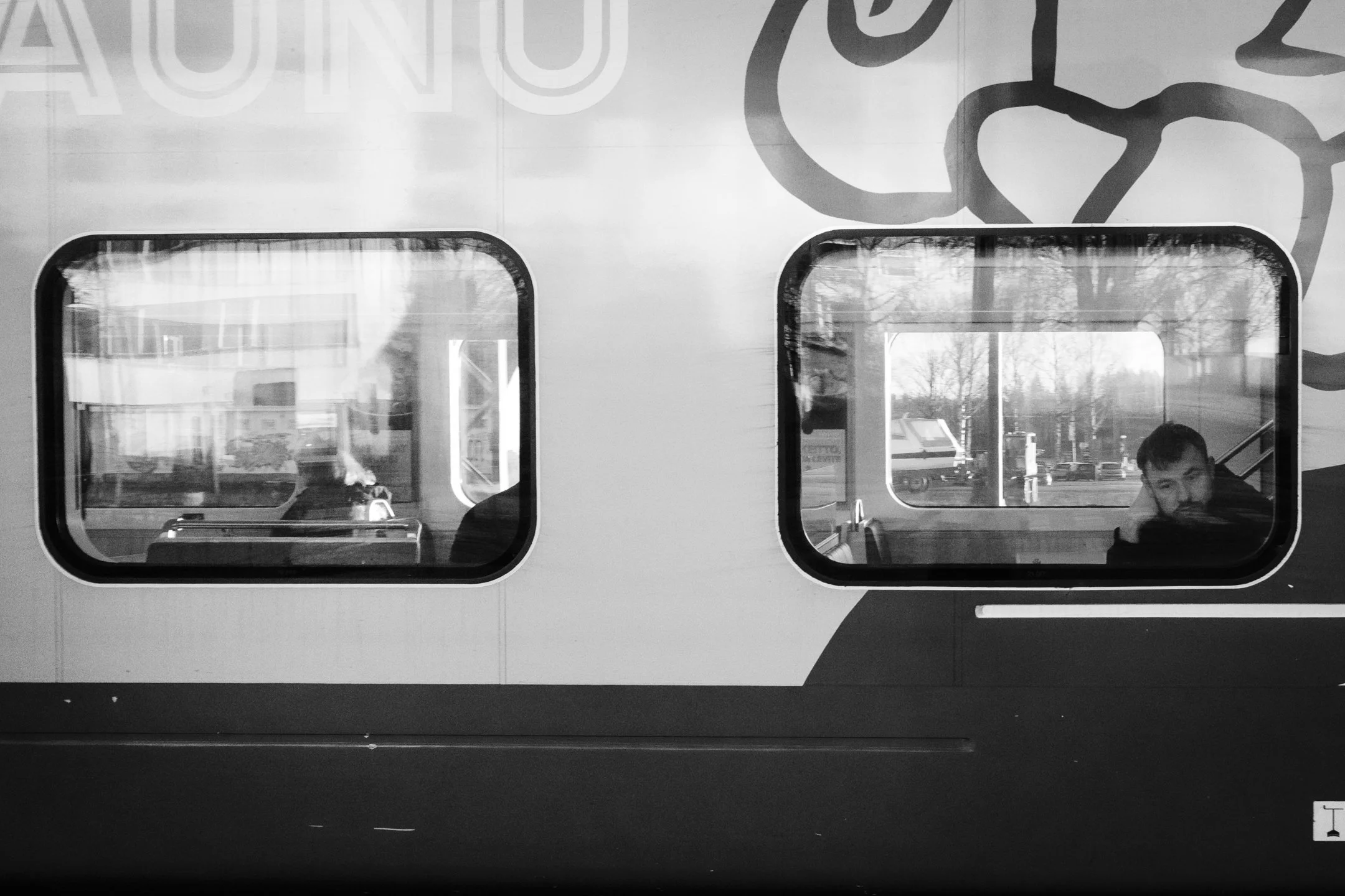 Kahden ihmisen heijastukset junan ikkunassa, sisällä istuvat ja taustalla ulkoilma näkyy.