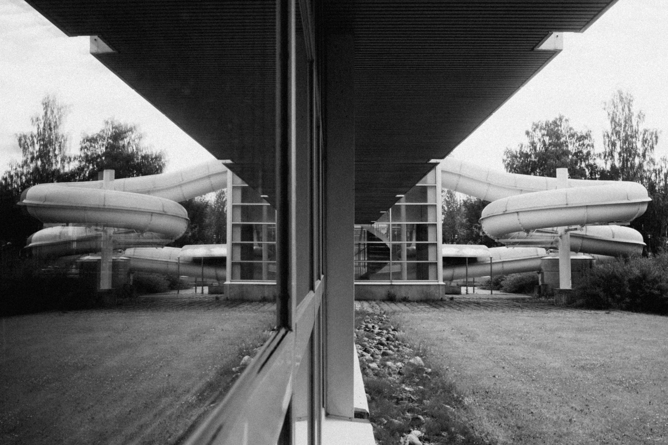 Kuvassa on vesiliukumäki ja rakennus, jonka edessä on ikkuna ja peilikuva siitä, maisema on taustalla, kaikki mustavalkoisena.